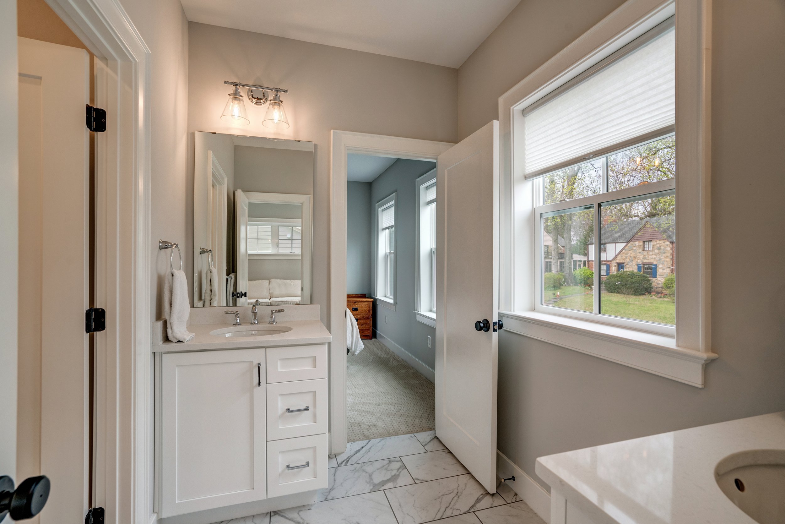Bathroom with a small white vanity, mirror, and large window showing a house and trees outside.