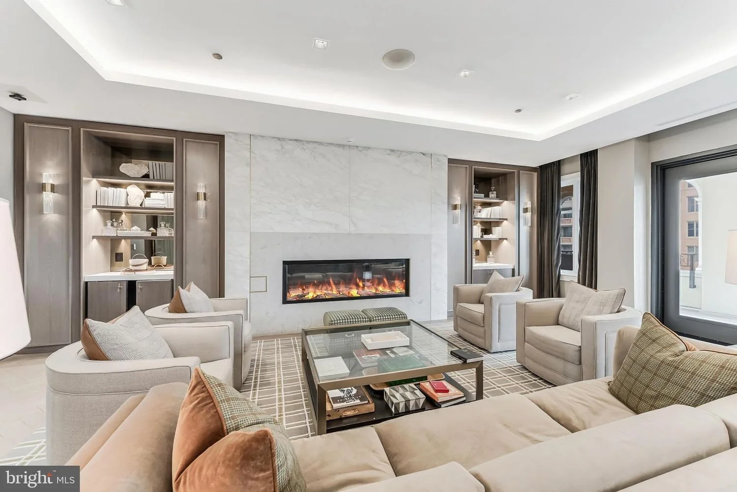 Modern living room with beige sectional sofa, armchairs, a glass coffee table, a fireplace with a marble surround, and built-in shelves with decor, large windows with black curtains