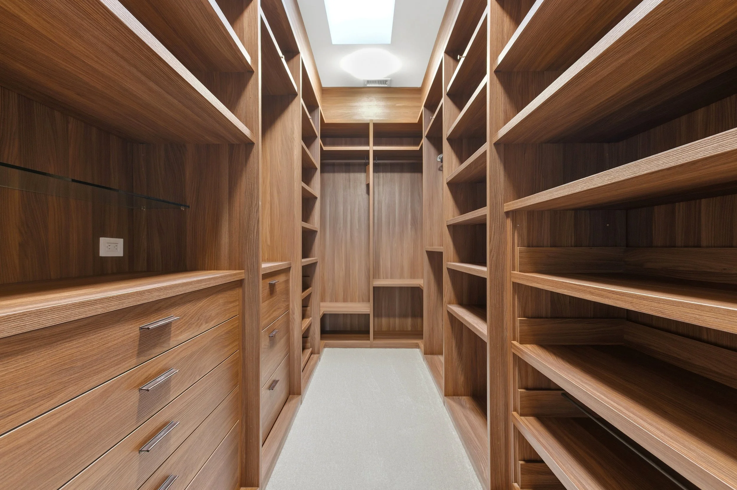 Empty walk-in closet with wooden shelving and drawers on both sides, beige carpet, and ceiling light.