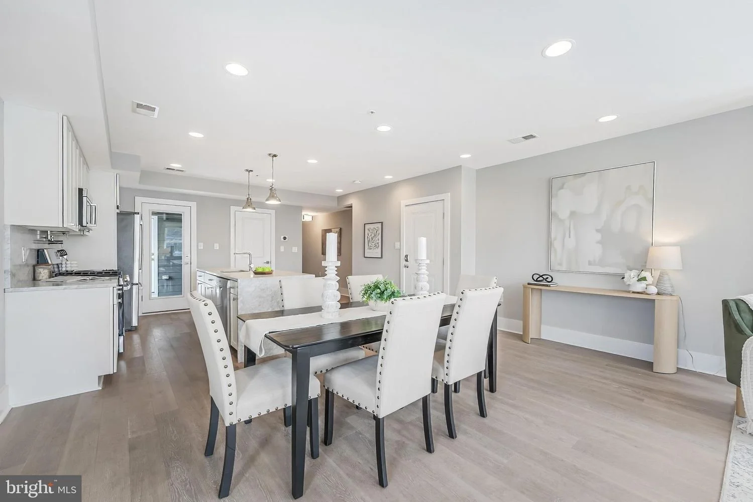 Open-concept kitchen and dining area with white walls, light wood flooring, a dark wood dining table with white upholstered chairs, a kitchen island with bar stools, and modern decor including candles and a framed art piece.