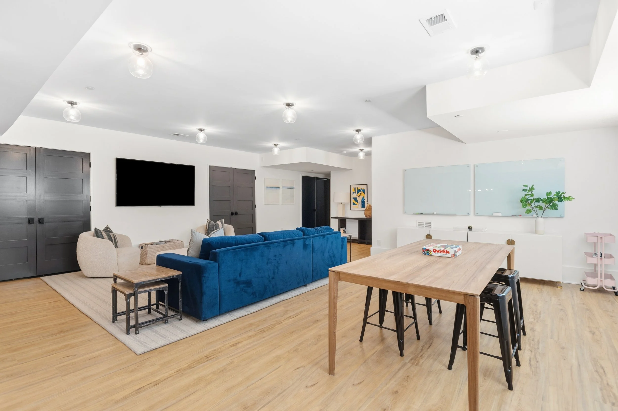 A modern living room with white walls and ceiling, hardwood floors, and various furniture including a beige armchair, a blue velvet sofa, small tables, and a wooden dining table with black stools. There are closed gray doors, framed artwork, a whiteb