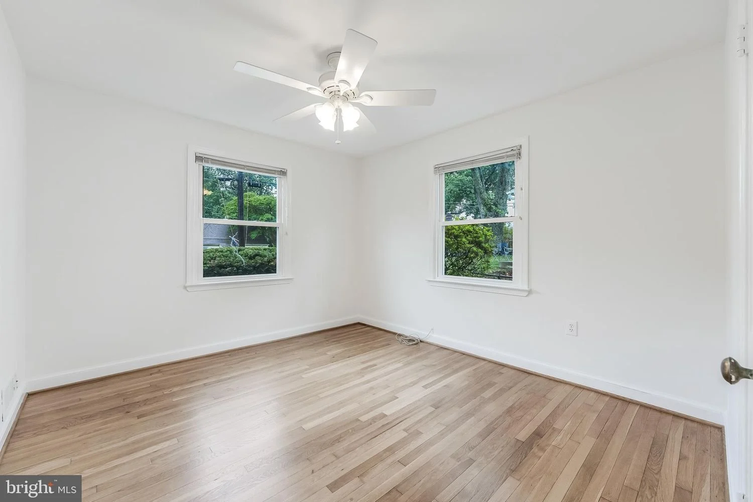 Empty room with hardwood flooring, white walls, two windows with blinds, and a ceiling fan with lights