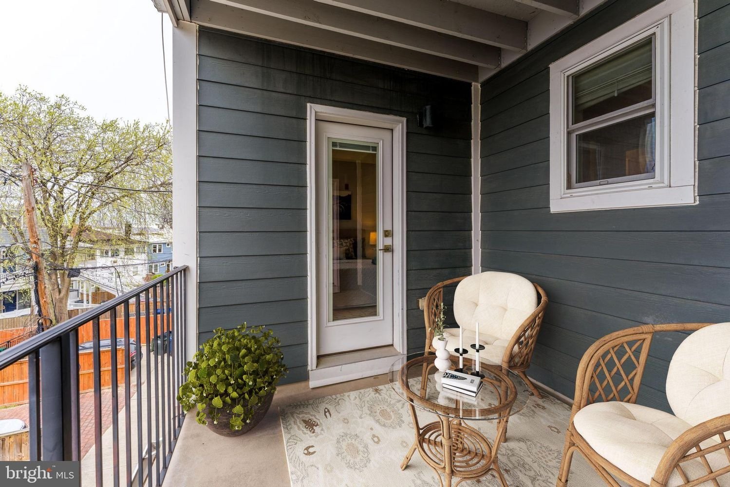 Outdoor balcony with two rattan chairs with cream cushions, a small glass-top table with candles, a potted plant in a brown pot, and a beige area rug. The balcony has gray wooden siding, a white door, and window, with a view of some trees and neighbo