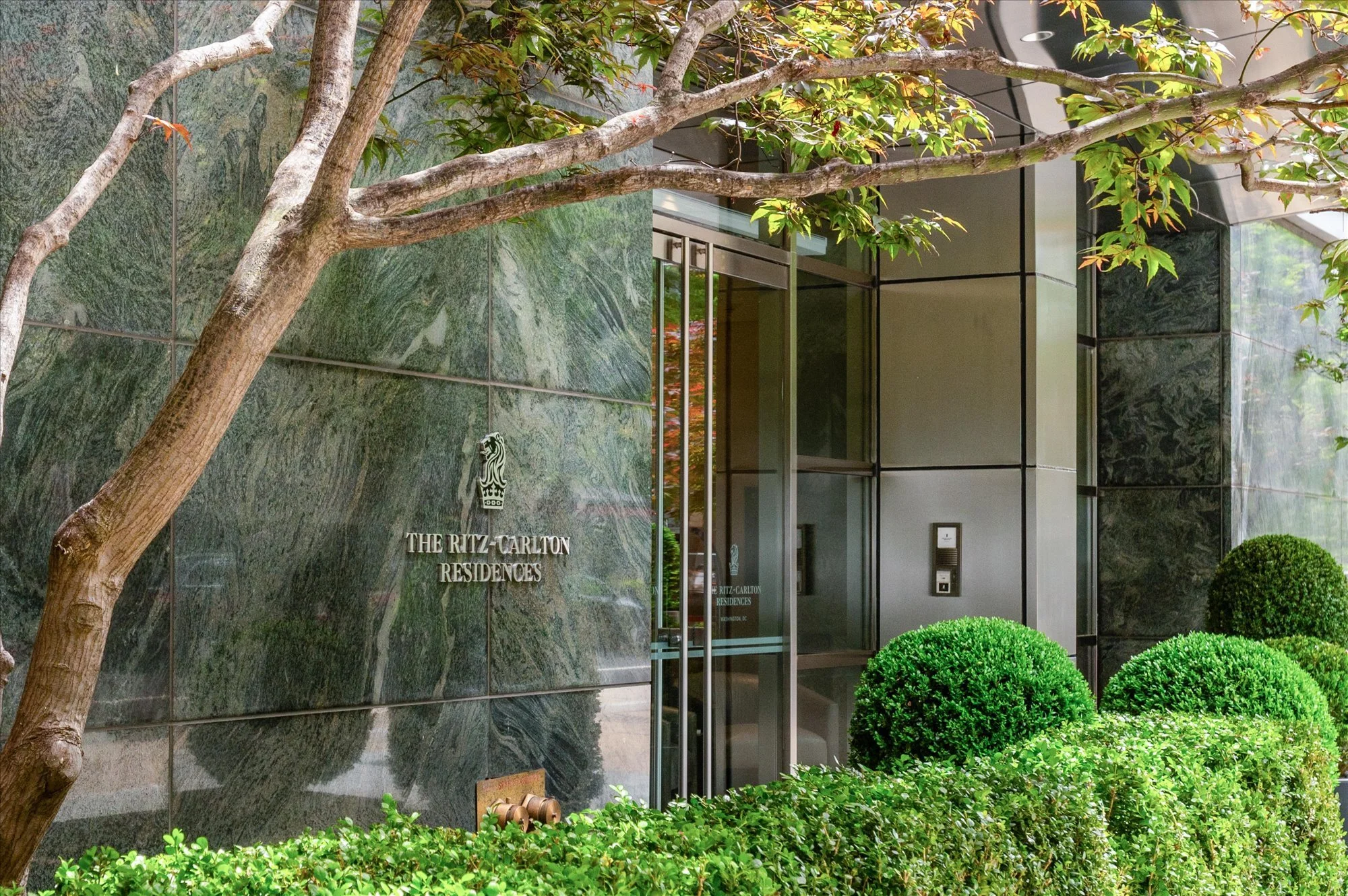 Exterior view of The Ritz-Carlton Residences with green foliage and trees, glass entrance doors, and a marble wall.