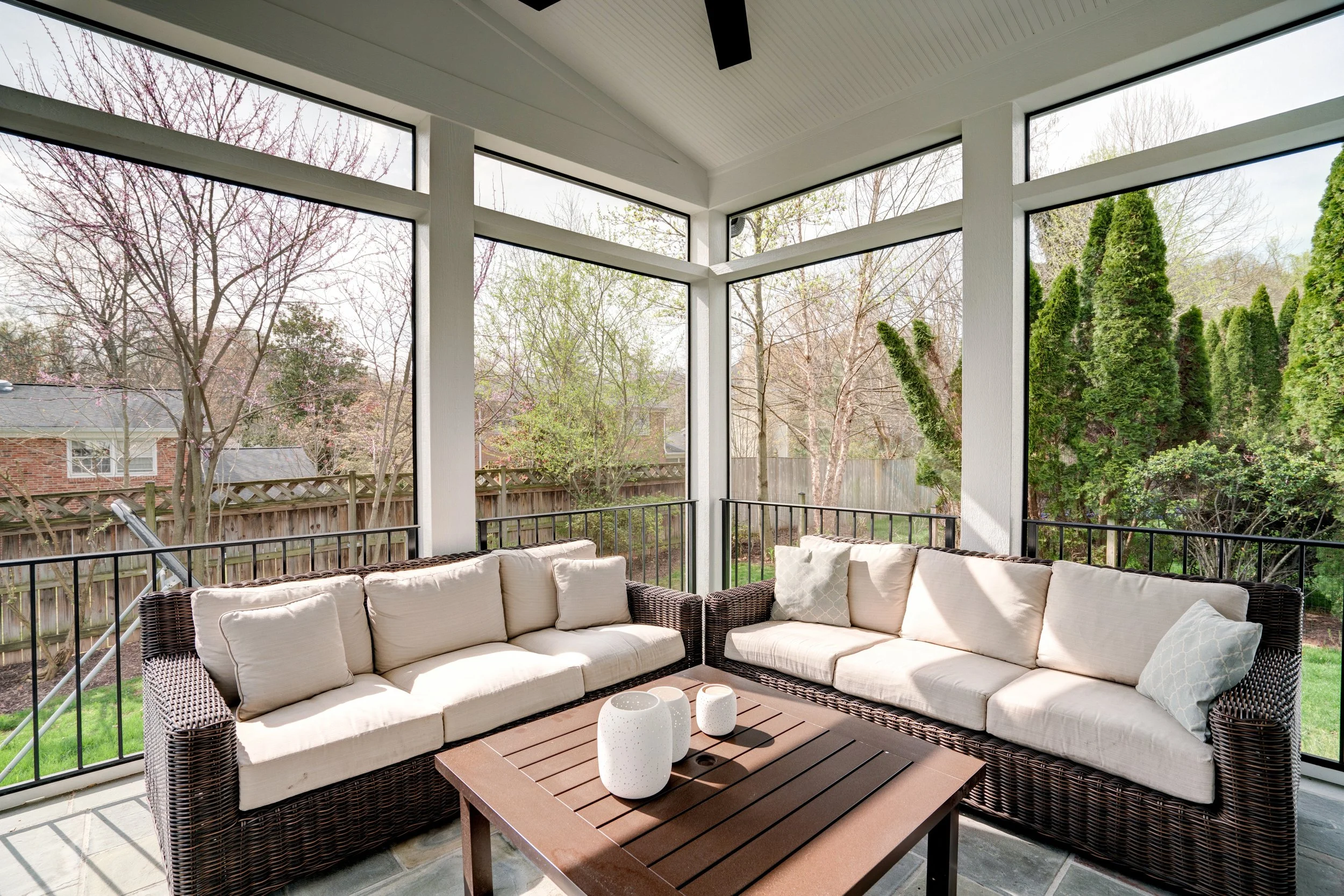 Enclosed porch with wicker cushioned seating, a wooden coffee table, and large windows showing trees and neighboring houses outside.