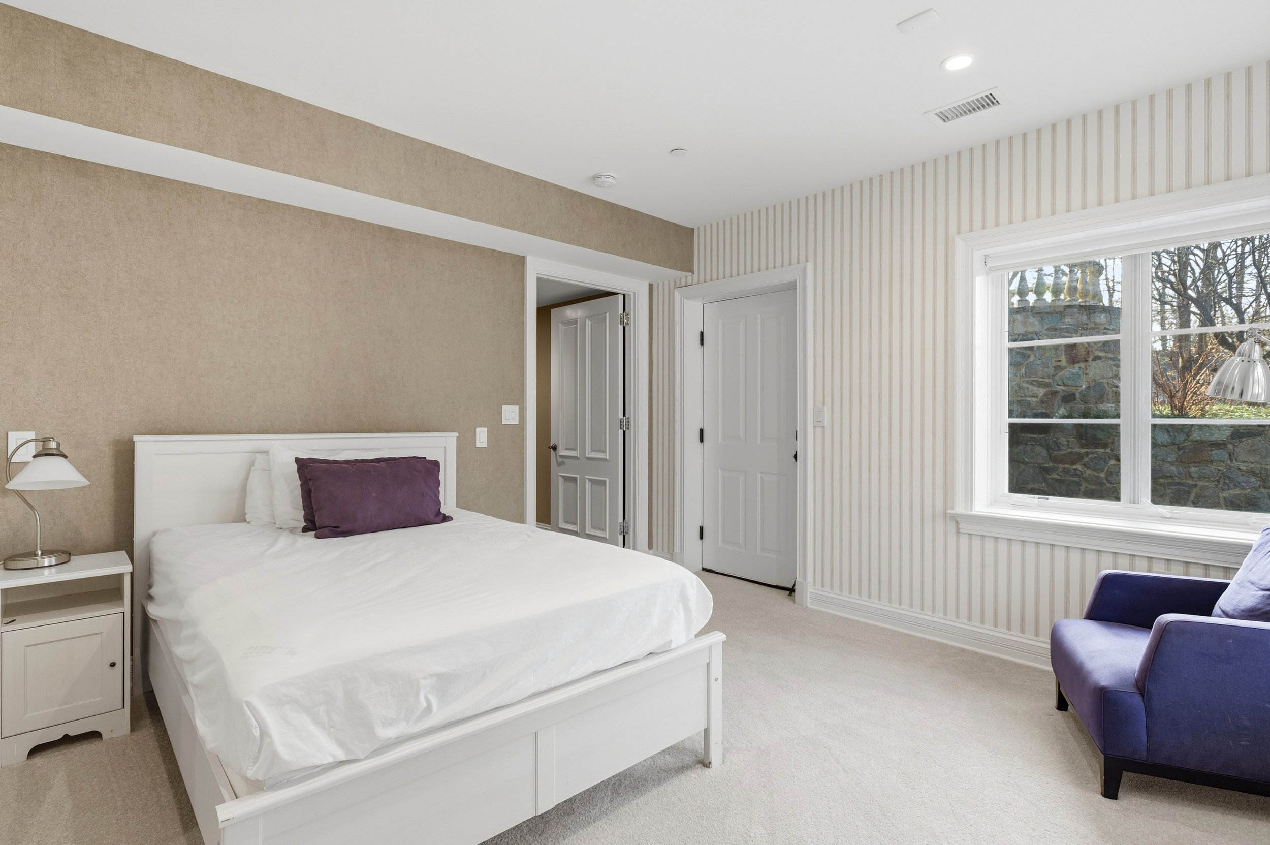 Bedroom with white bed, purple pillow, side table and lamp, beige and striped wallpaper, large window with view of trees and stone wall.
