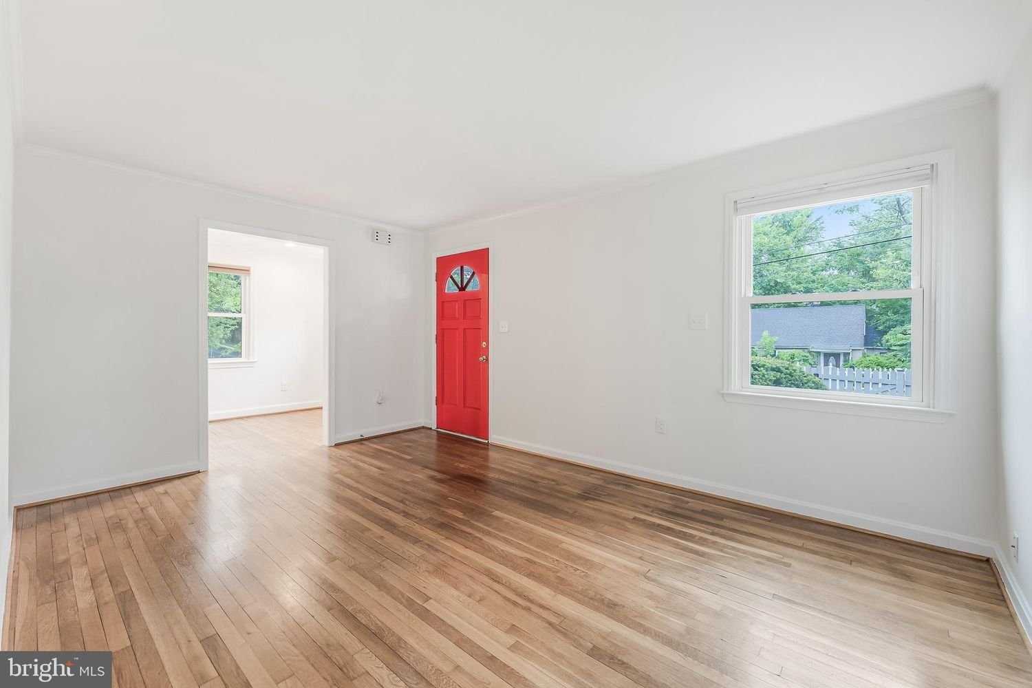 Empty living room with hardwood floors, white walls, a red front door, and large windows showing greenery outside.