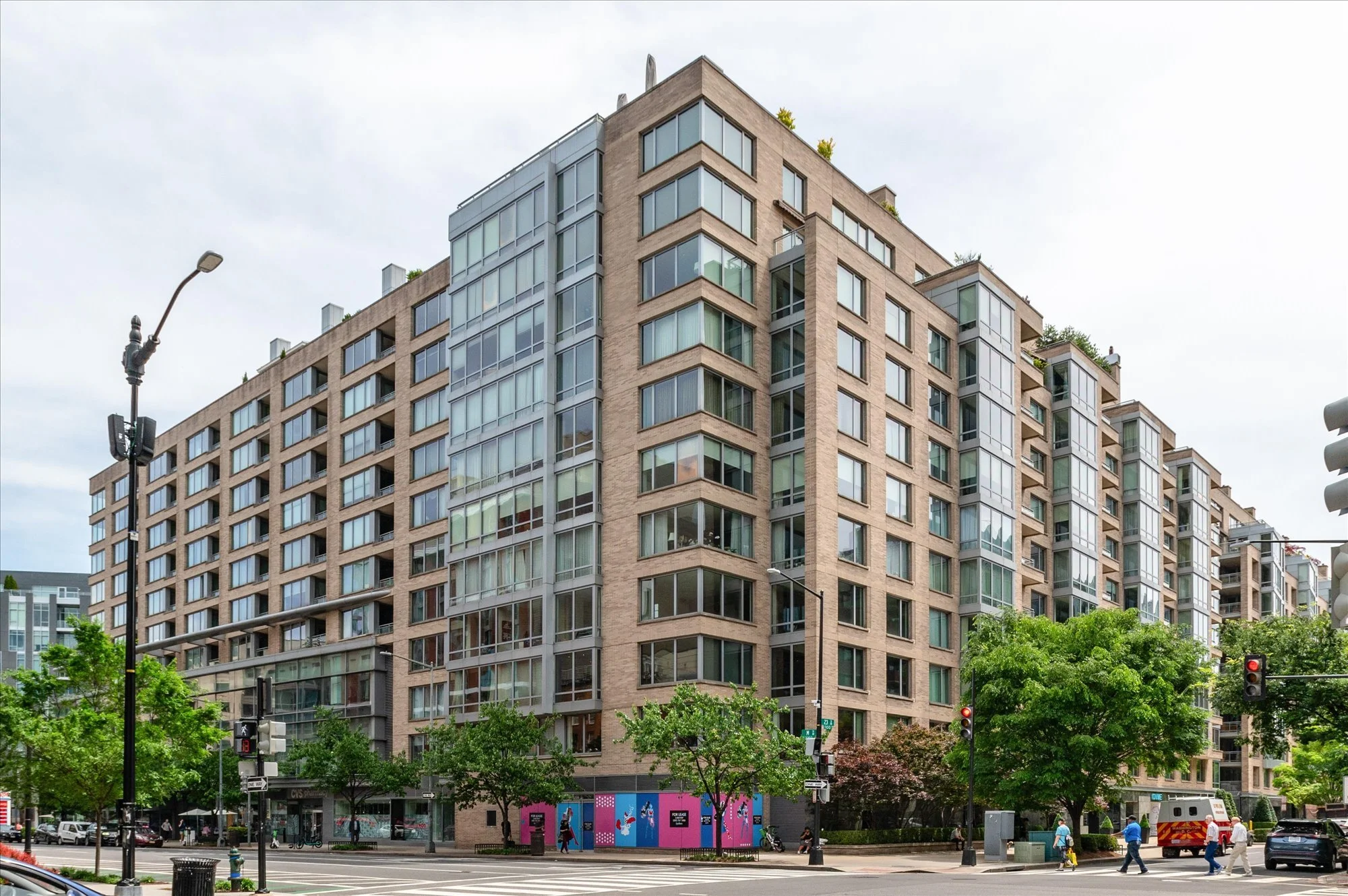 A tall modern residential building with large glass windows and balconies, situated at a city street corner with busy traffic lights and pedestrians walking on the sidewalk.