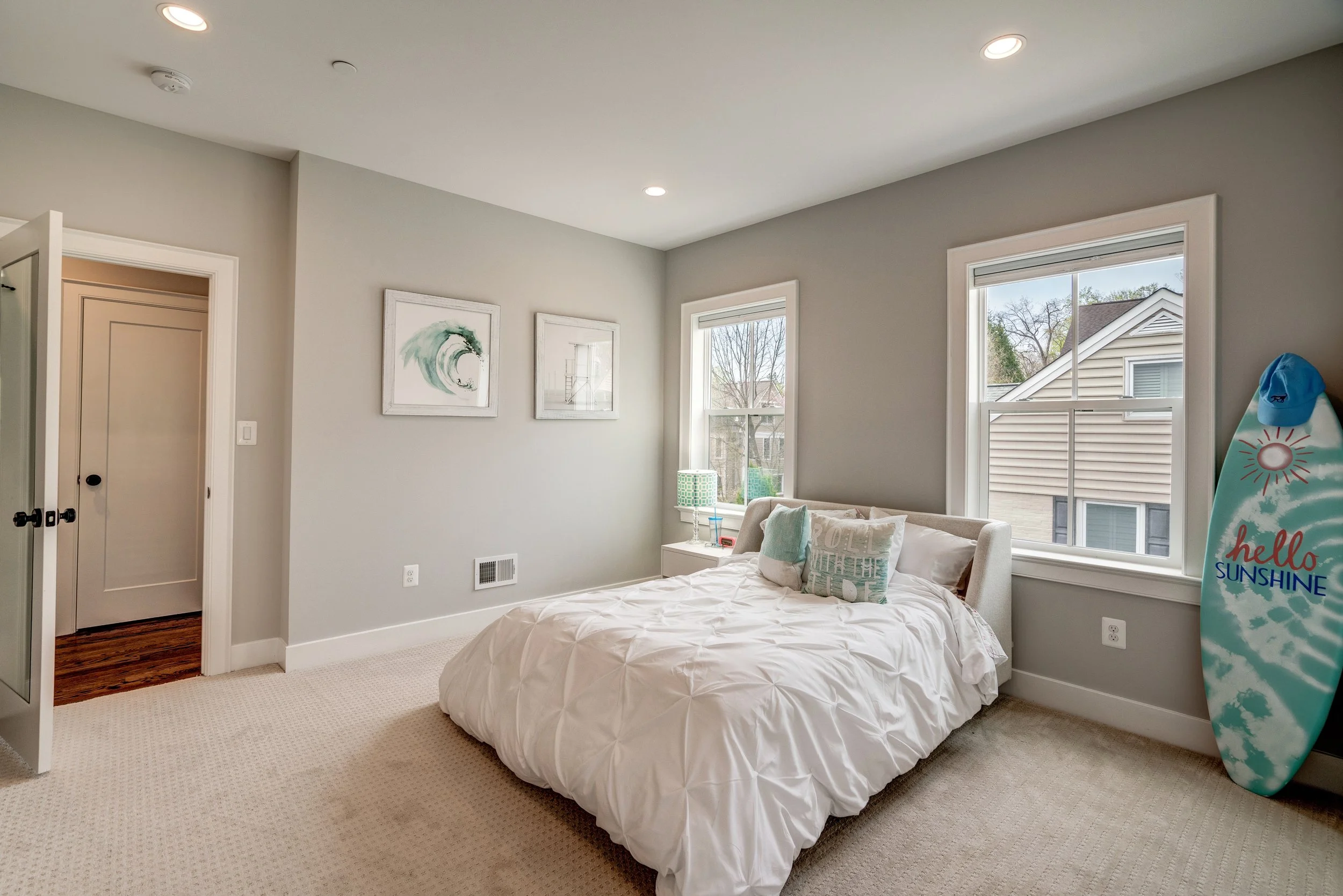 A bedroom with a bed covered in a white comforter, decorative pillows, two windows with blinds, a nightstand with a lamp, framed artwork on the wall, and a surfboard with a blue cap that says "hello sunshine".