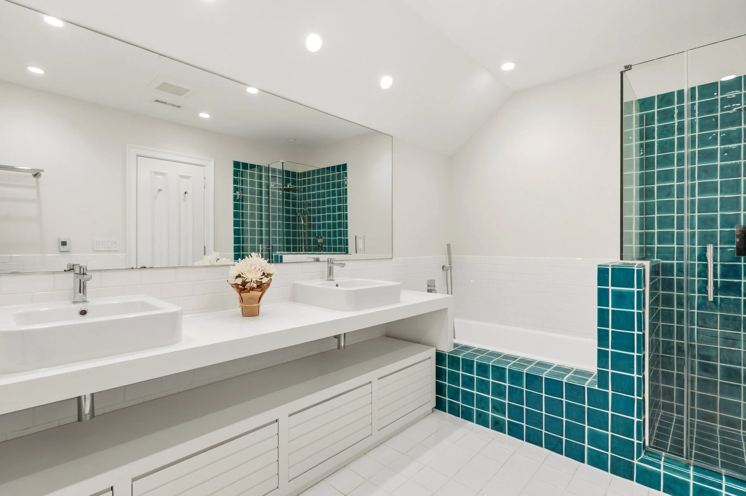 Modern bathroom with dual white sinks, large mirror, turquoise tiled shower, white walls, and white floor.