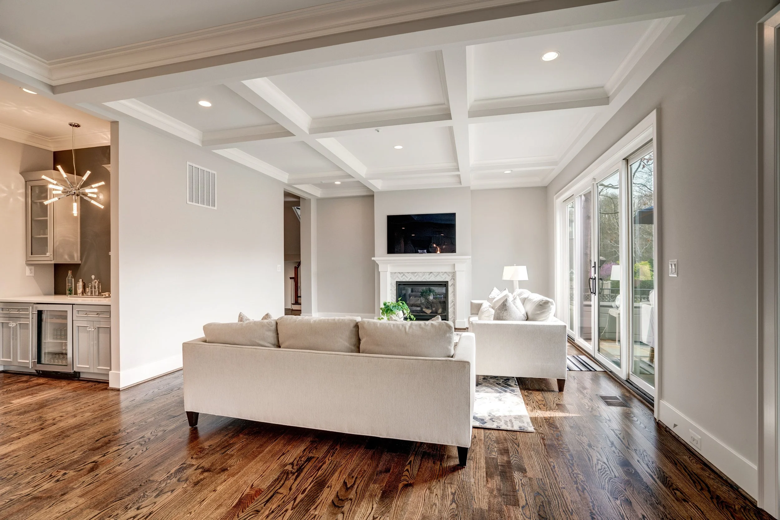 Bright living room with white ceiling beams, recessed lighting, large windows, white sofa, and fireplace with mounted TV.