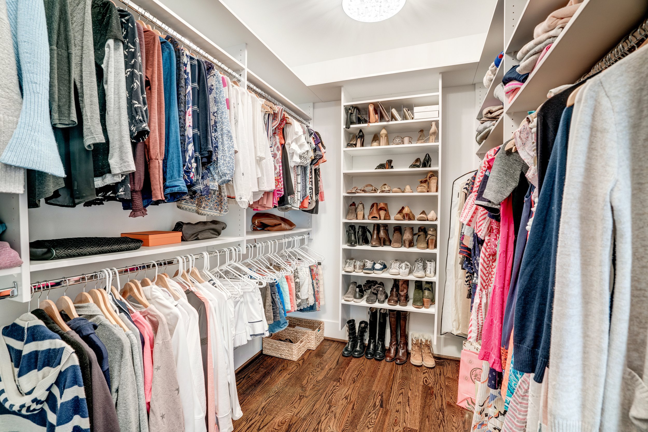 A walk-in closet with white shelving and hanging clothes, including shirts, dresses, and jackets, along with shelves of shoes and boots, and a wooden floor.