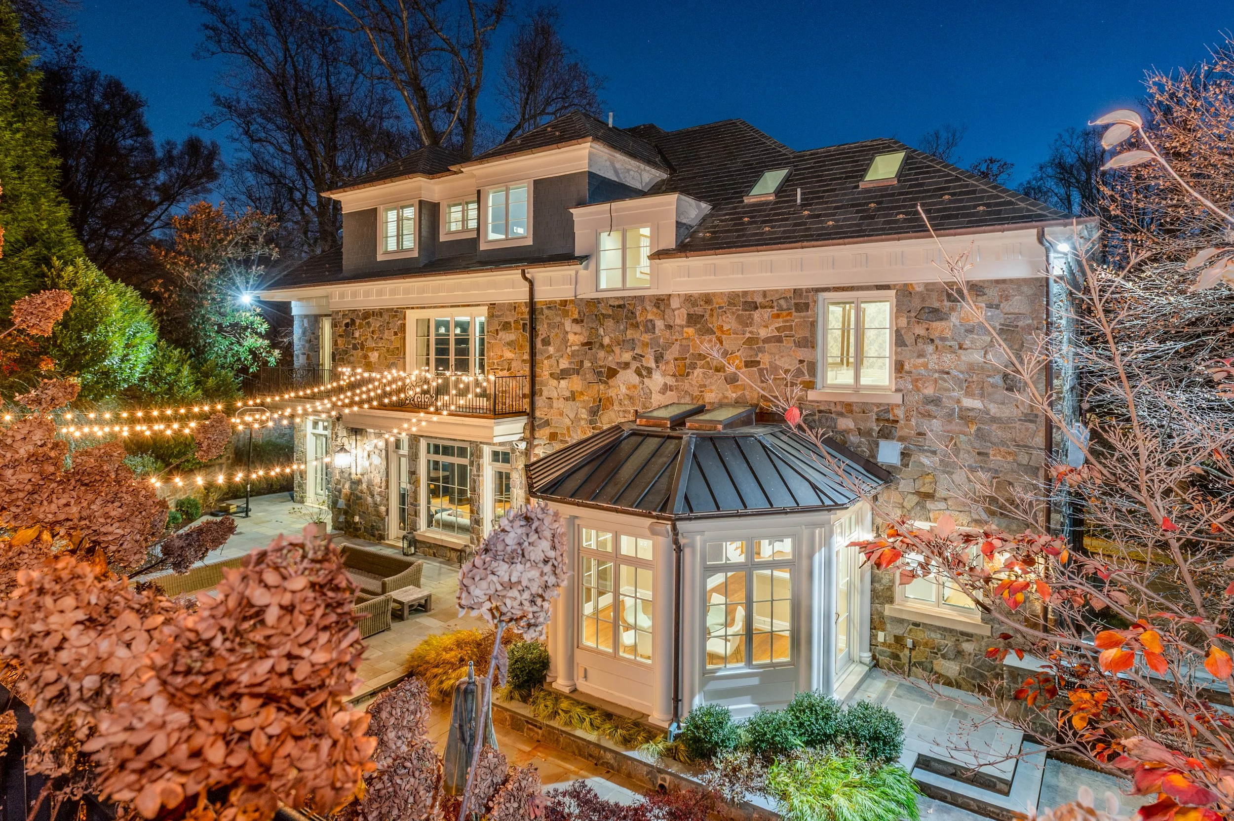 Night view of a large house with stone and white siding, illuminated windows, and string lights on a patio. Trees with autumn-colored leaves surround the house.