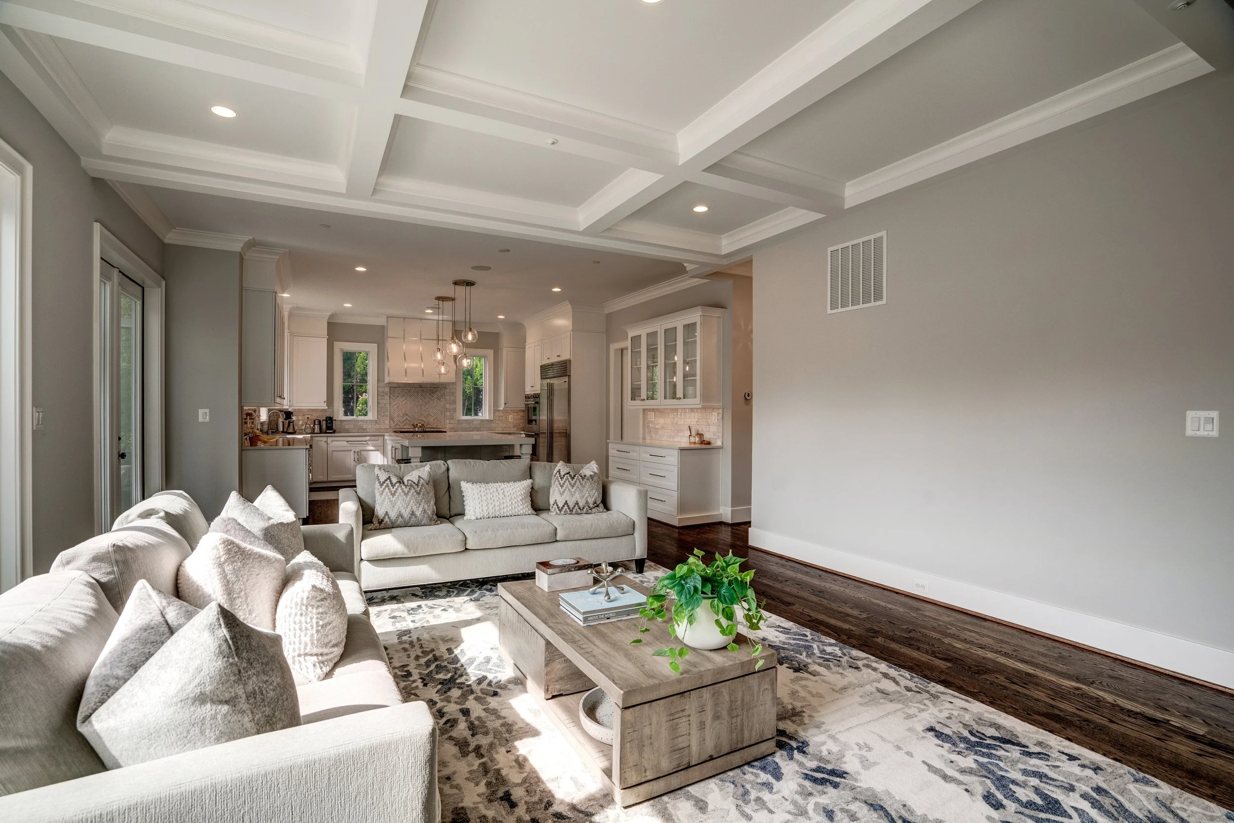 Open-concept living room and kitchen with beige sofas, a wooden coffee table, kitchen cabinetry, and pendant lighting in a modern home.
