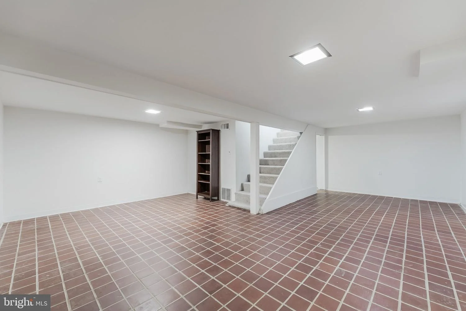 Empty basement with white walls, brown tiled floor, staircase with gray carpet, and a small dark wooden bookshelf.