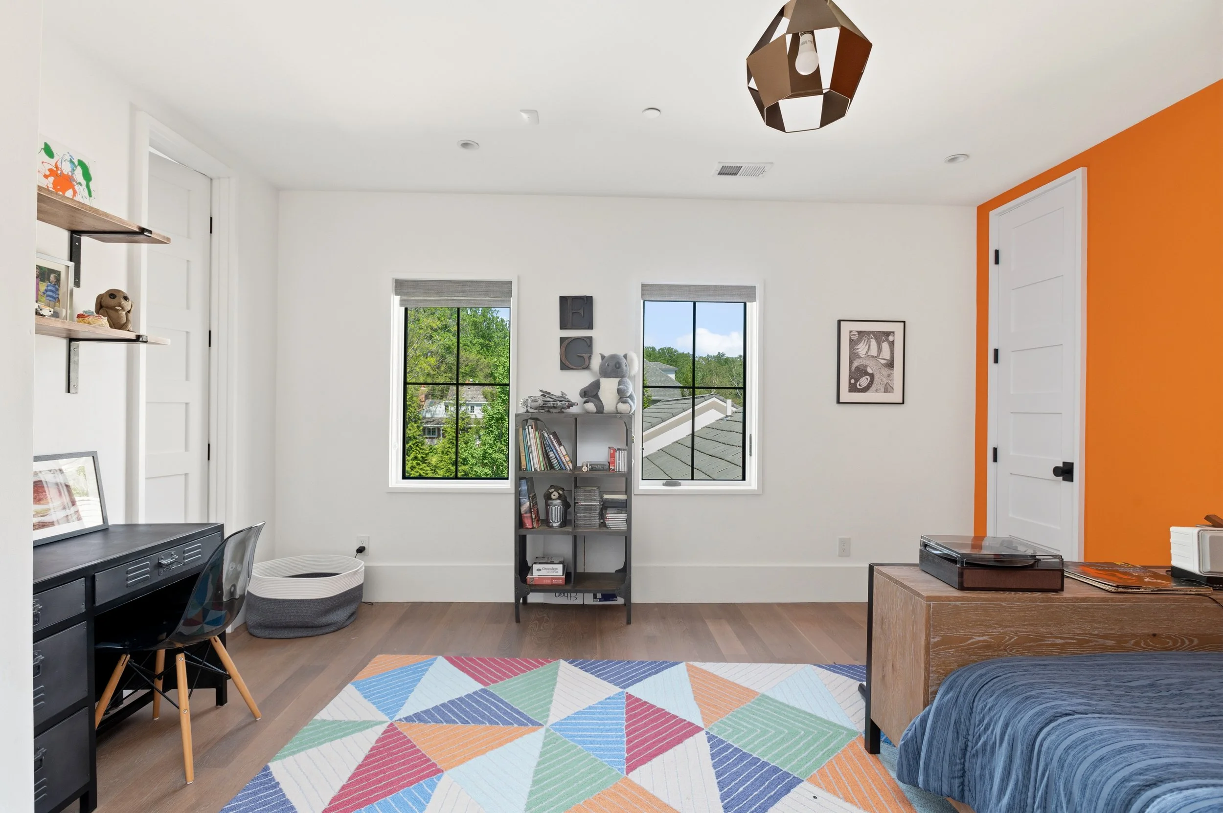 View of a bright modern room with two large windows, colorful geometric rug, and a mix of furniture including a black desk, a gray and white pet bed, a multicolored chair, a bookshelf with books and a plush koala bear, and a wooden bench. One wall is