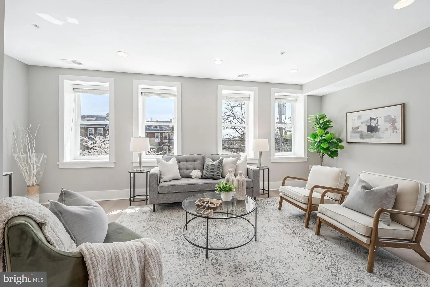 Bright living room with light gray walls, three large windows, and modern furniture, including a gray sofa, white armchairs, and a round coffee table with decorative vases and plants.