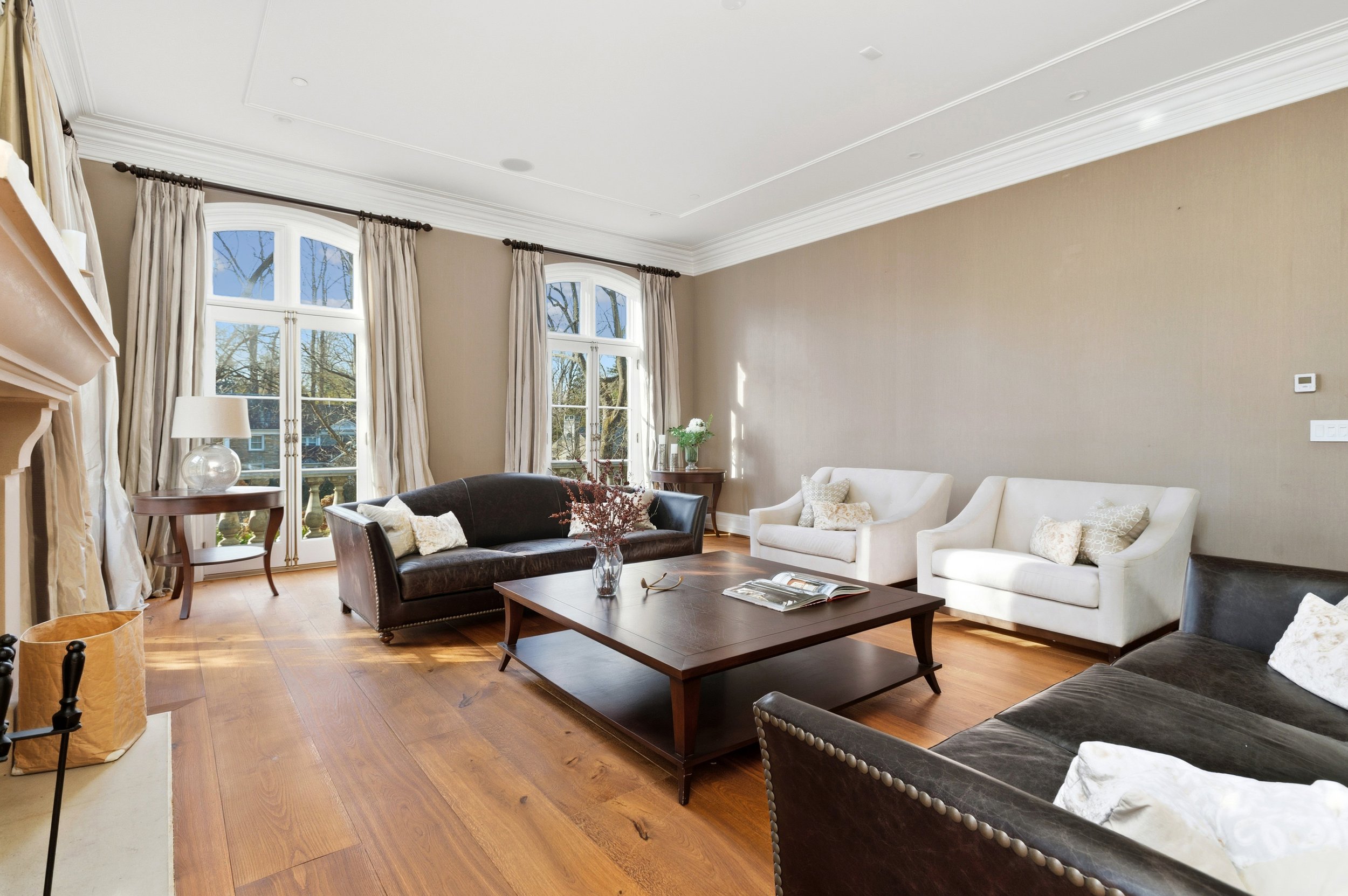 Living room with large windows, beige walls, wooden floors, and various sofas and chairs, decorated with cushions, a coffee table, and a vase with flowers.