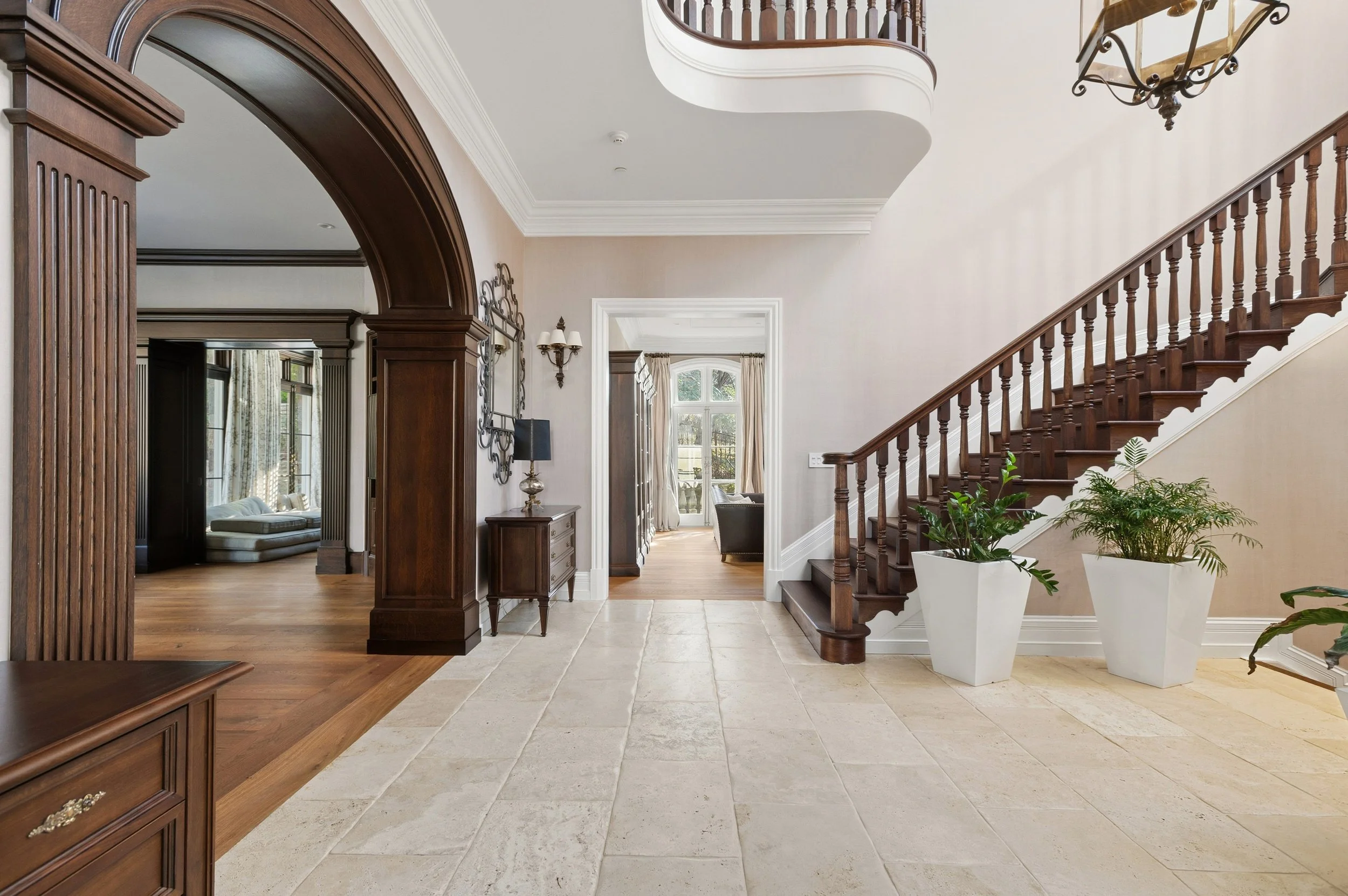 Interior of a house foyer with tiled floor, wooden staircase with bannister, and a chandelier hanging from the ceiling. Visible part of a living room with a couch and large windows with curtains.