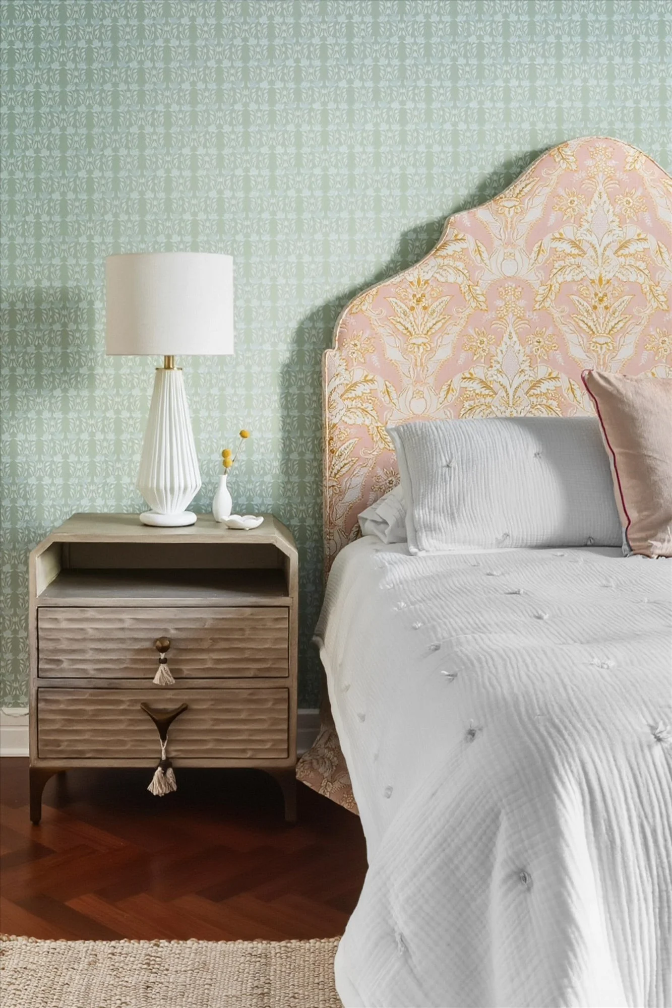 A bedroom with a pink and gold patterned headboard, white bedding, and a nightstand with a white table lamp and small decorative items. The wall has a light green wallpaper with subtle floral motifs, and there is a beige woven rug on the wooden floor