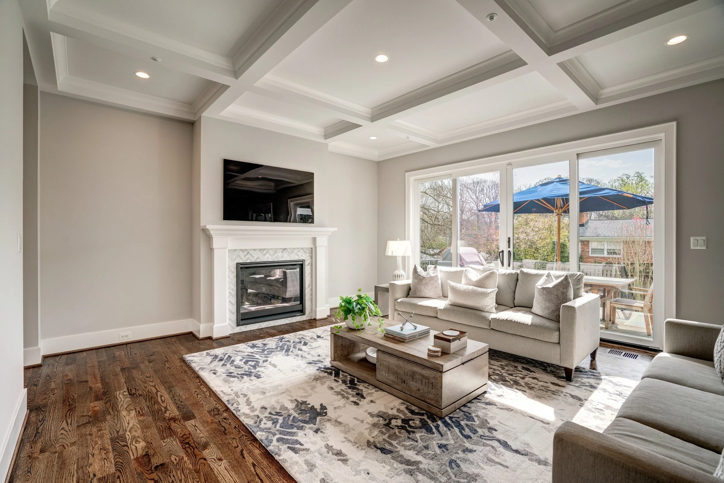 Living room with large sliding glass door, white couch, coffee table, fireplace, flat-screen TV, area rug, and outdoor patio with blue umbrella