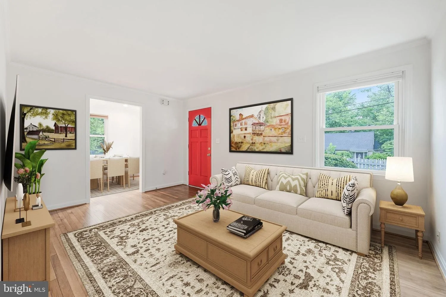 Living room with white walls, a beige sofa with pillows, wooden coffee table with flowers, window showing greenery outside, and a wall art painting. There's a side table with a lamp, and a doorway leading to a dining area with a table and chairs. A r