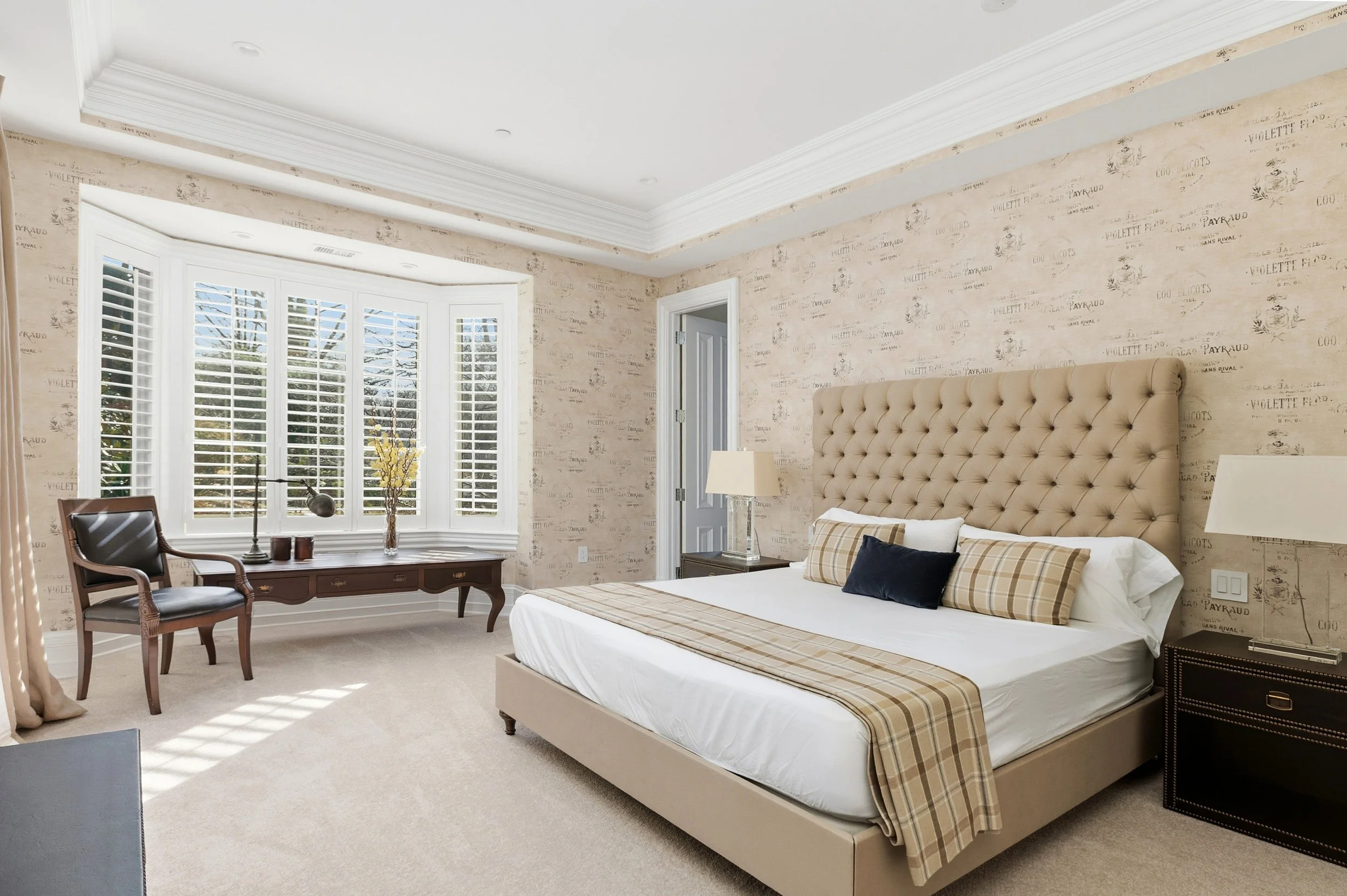 A bedroom featuring a large bed with plush beige headboard, plaid and solid pillows, a wooden desk with a desk lamp and decorative items, a chair near the window, and beige carpet flooring with window shutters.