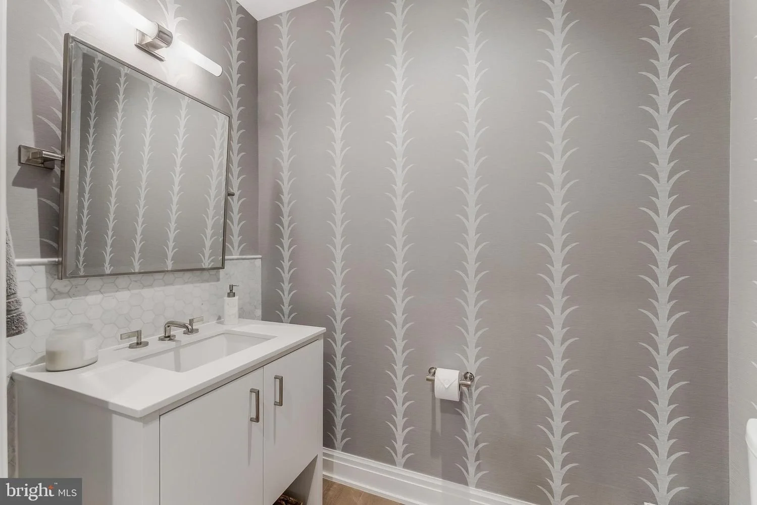 Modern bathroom with gray wallpaper featuring a leaf pattern, a white vanity with a sink, mirror, and decorative accessories, and a toilet paper holder on the wall.