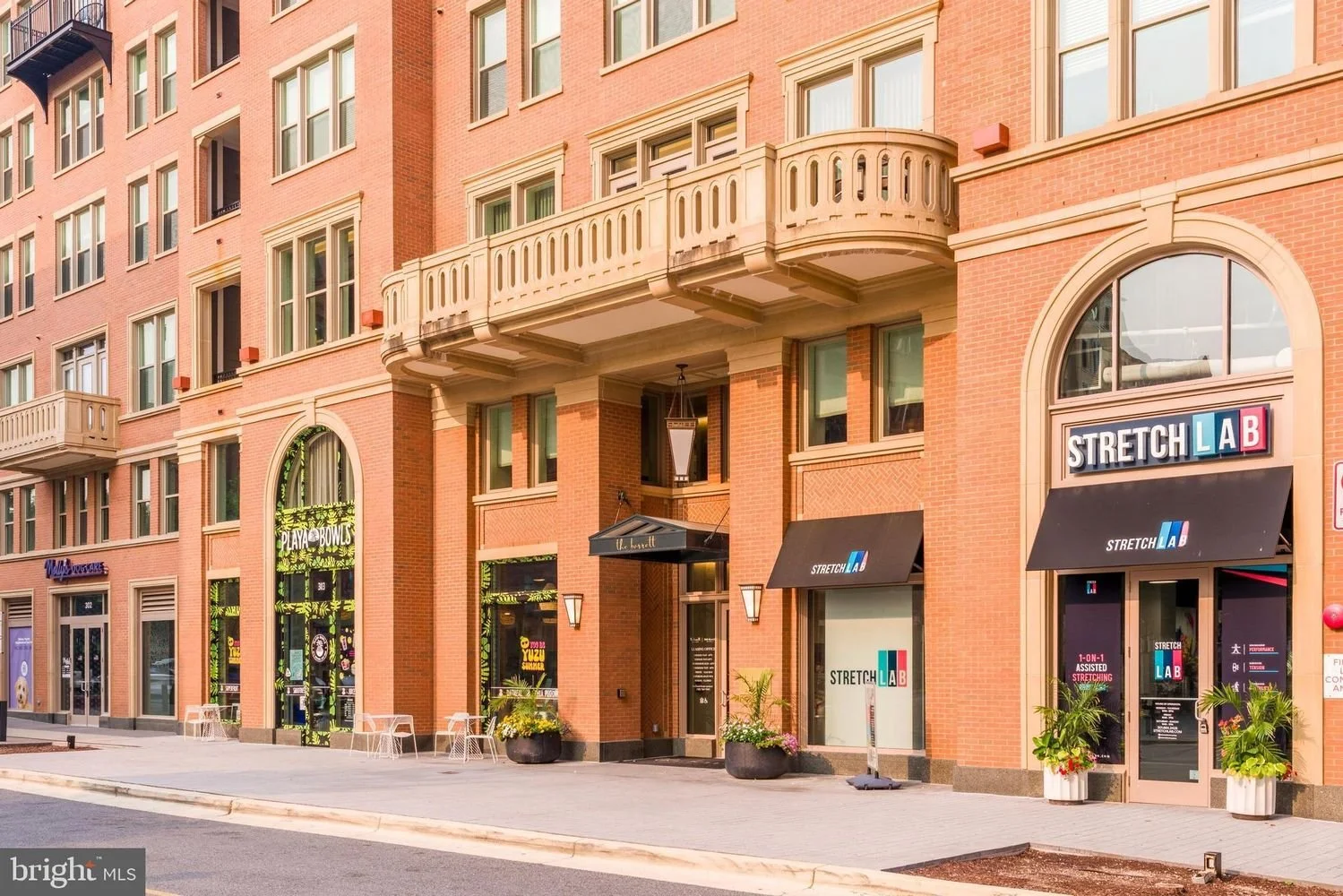 Street view of a brick building with various businesses, including Stretch Lab and Playa Bowls, featuring outdoor seating and potted plants.