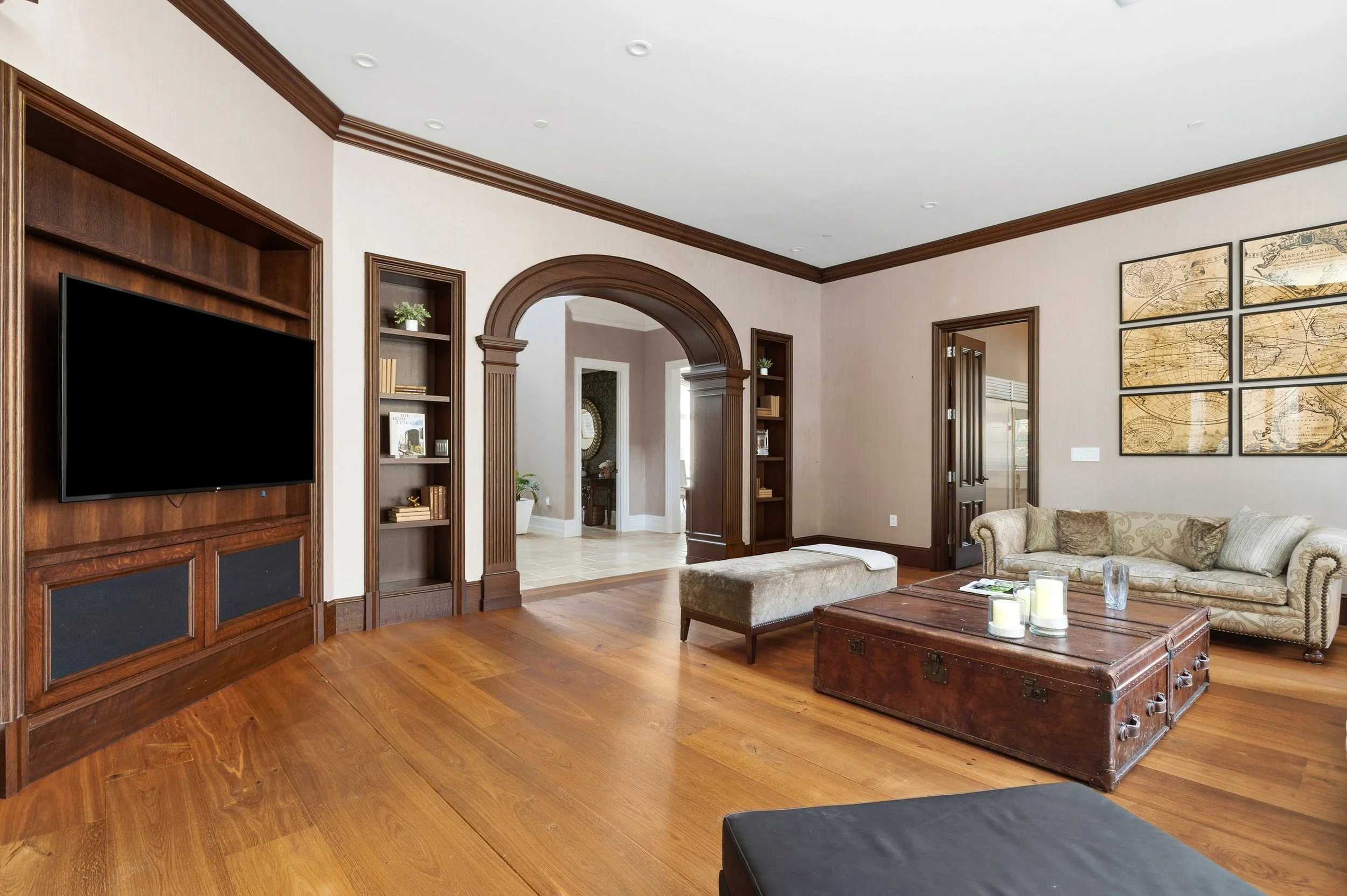Living room with hardwood floors, built-in wooden shelves, a large flat-screen TV, and a beige sofa with decorative pillows. There is a wooden trunk-style coffee table with candles on top, and an archway leading to other rooms. The walls are light-co