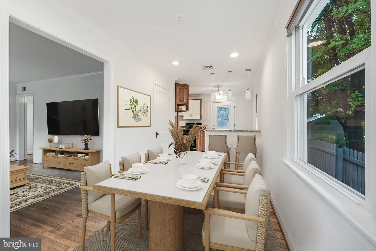 Dining area with a white table set with white bowls and napkins, six beige chairs, and a decorative vase with pampas grass, adjacent to a kitchen with wooden and white cabinets, next to a large window showing greenery outside.