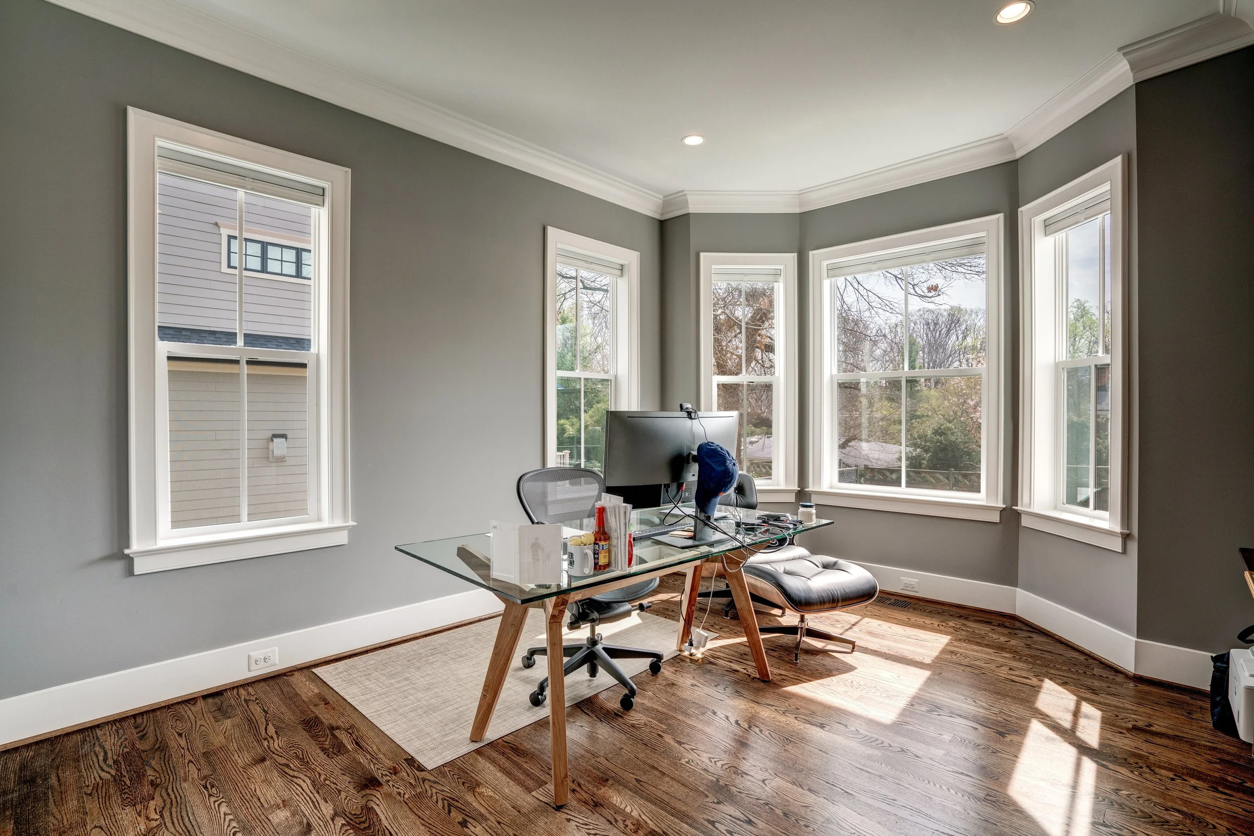 A home office with several large windows allowing natural light, a glass-top desk, an office chair, a monitor, and various small items including condiments and books, with hardwood floors and gray walls.