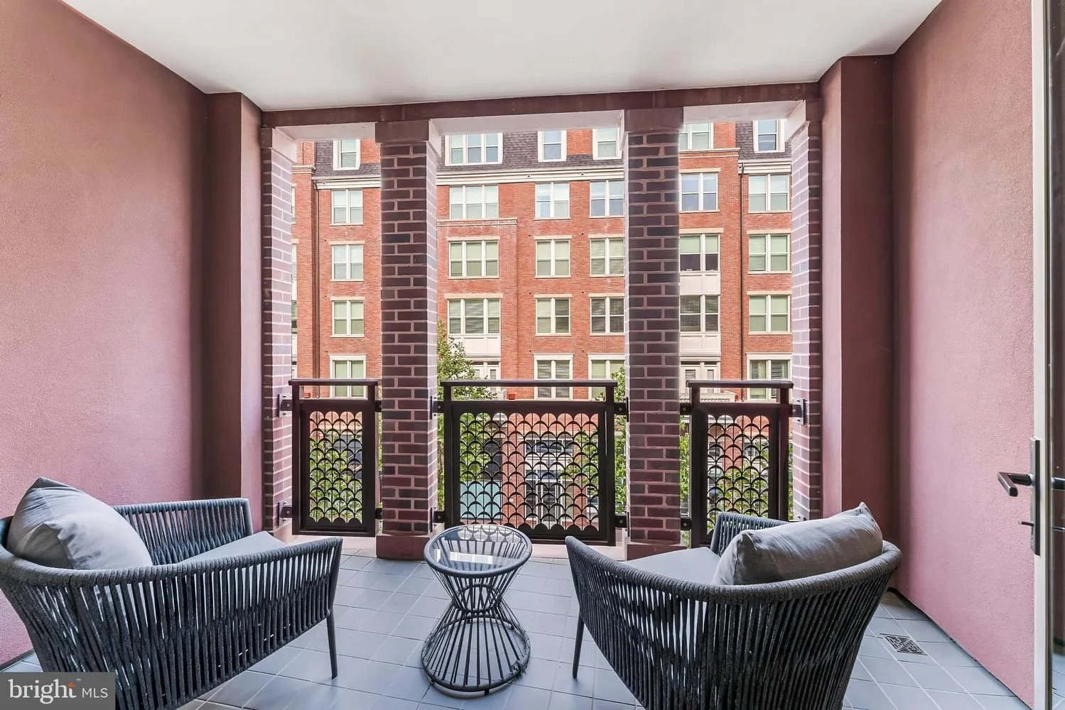Balcony with two black wicker chairs, a small round table, pink walls, brick columns, overlooking a neighboring red brick apartment building with multiple windows.
