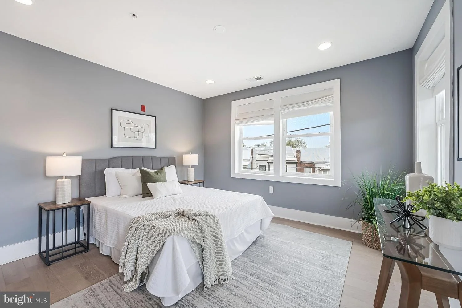 Bright bedroom with gray walls, white bedding, and two bedside lamps, a window with blinds, and modern decor including plants and a dresser.