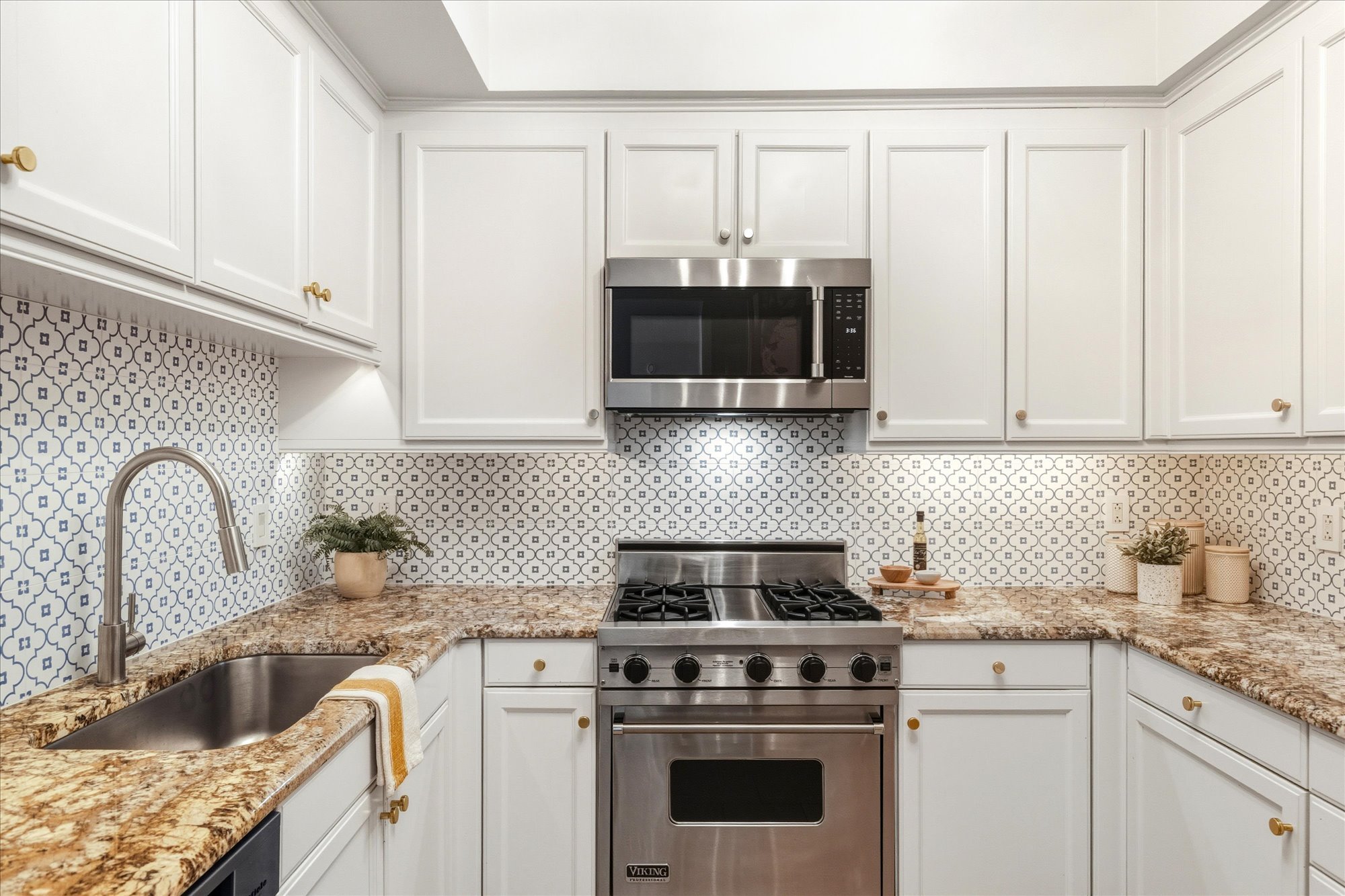 A modern kitchen with white cabinetry, a patterned backsplash, granite countertops, stainless steel appliances including a microwave and oven, a sink, and decorative plants on the counter.