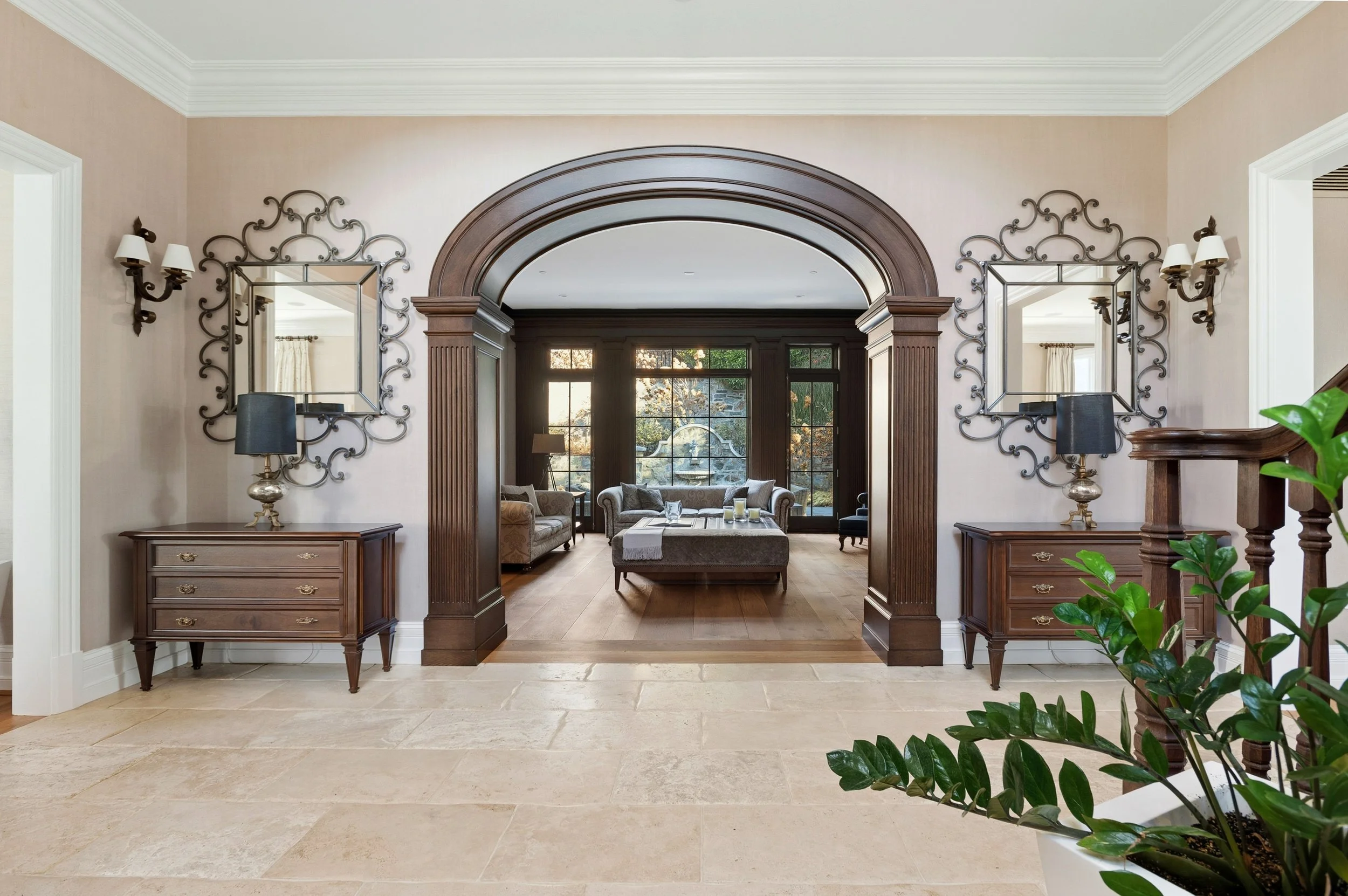 A spacious living room viewed through an archway with dark wood trim. The room features a large window with a view of trees, a sofa, armchairs, and a coffee table. The walls are decorated with two mirrors and wall sconces. There are two matching wood