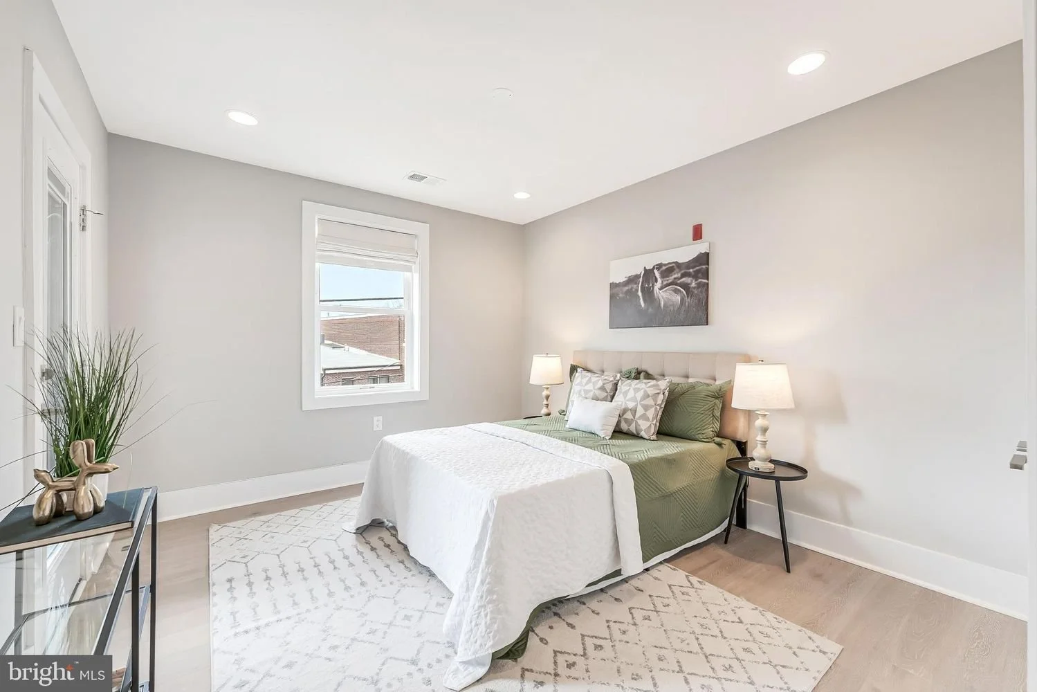 Modern bedroom with a bed, green and white bedding, nightstands with lamps, a window, a art piece of a horse, and decorative items on a black side table.