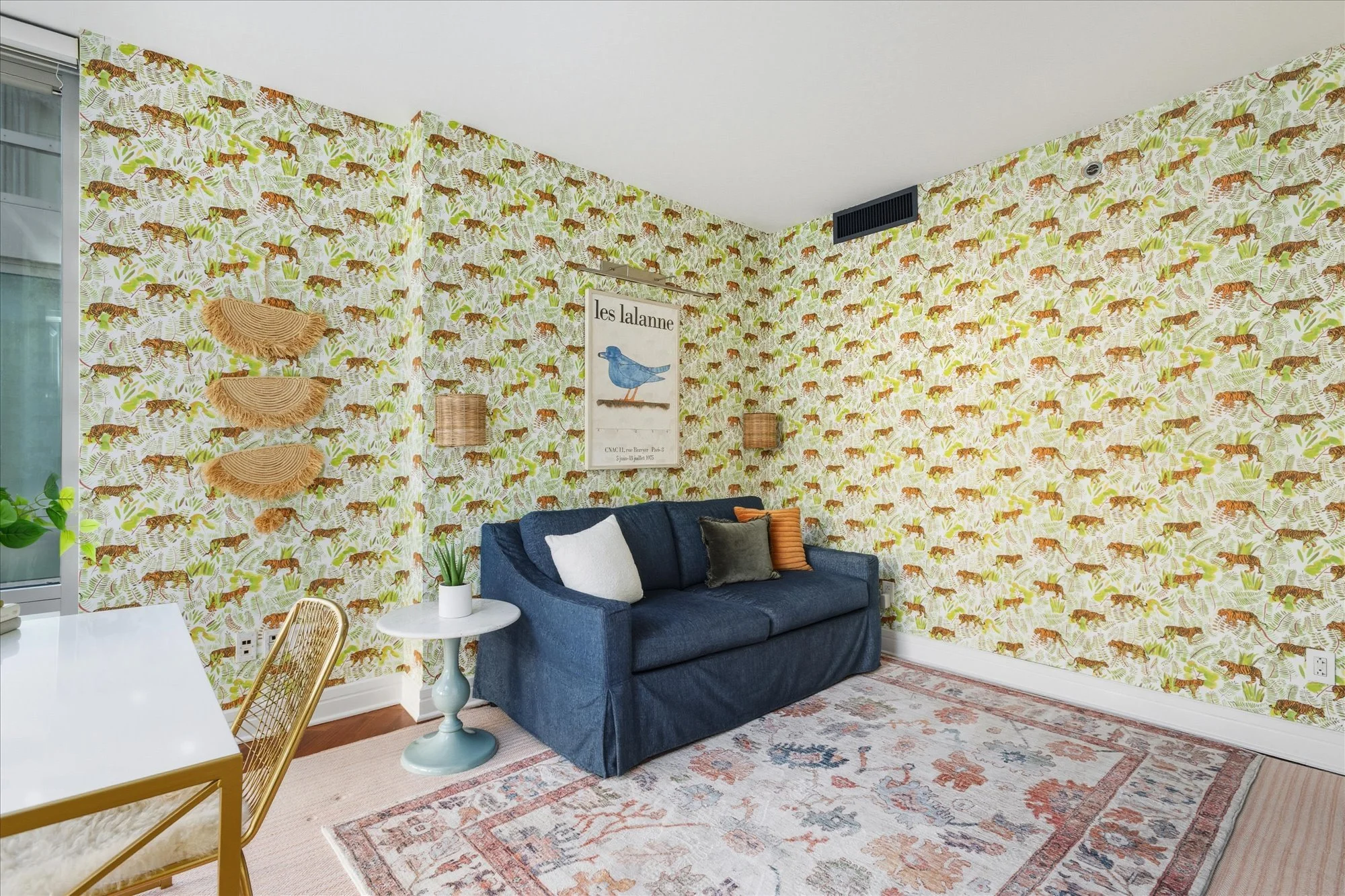A living room with tiger-patterned wallpaper, a blue sofa with pillows, a white side table with a plant, wall decorations including a framed picture of a bird, and a traditional rug on the polished wooden floor.