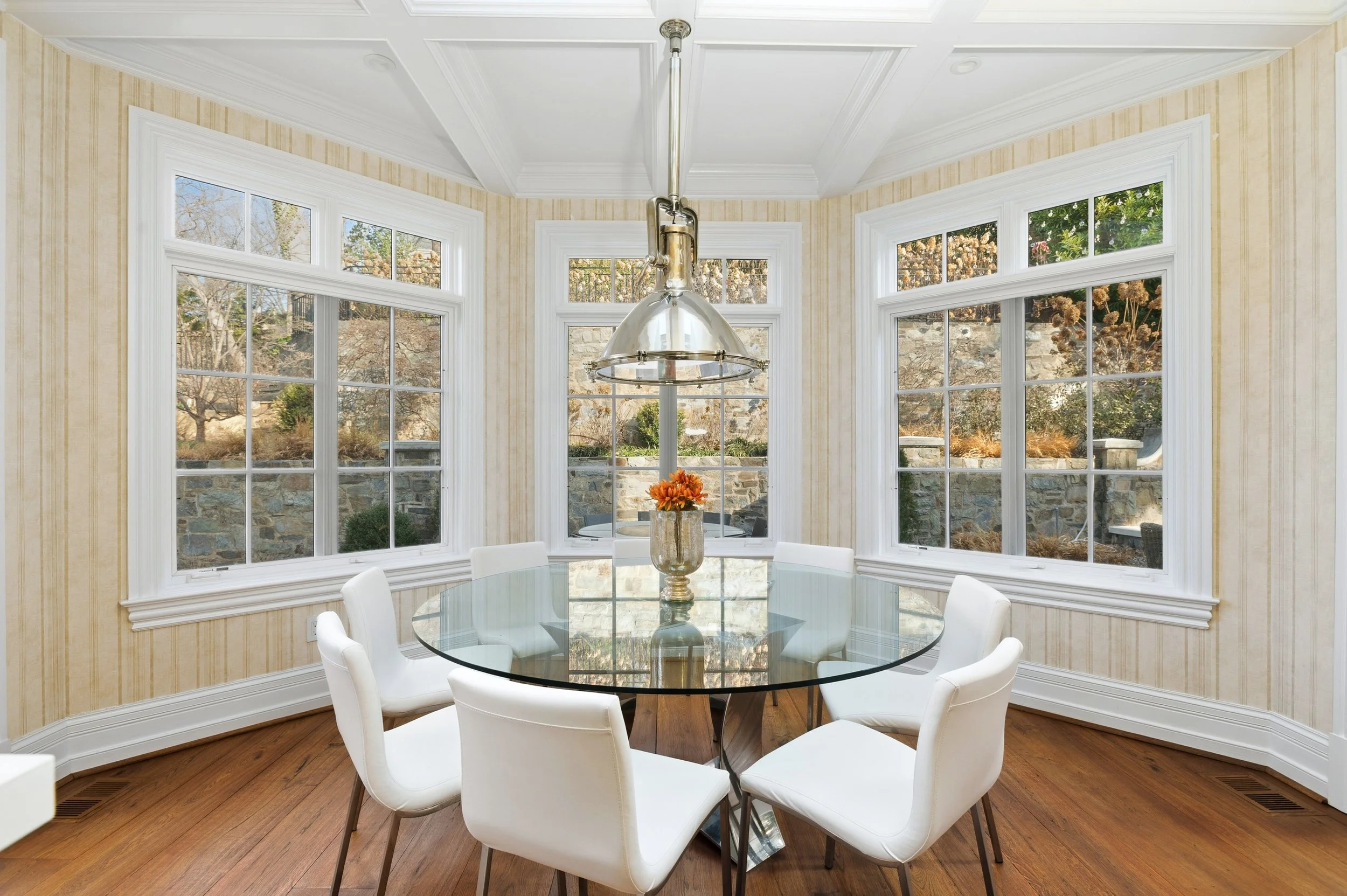 Bright dining area with large windows showing outdoor trees and shrubs, white chairs around a glass table, and a modern silver pendant light fixture.