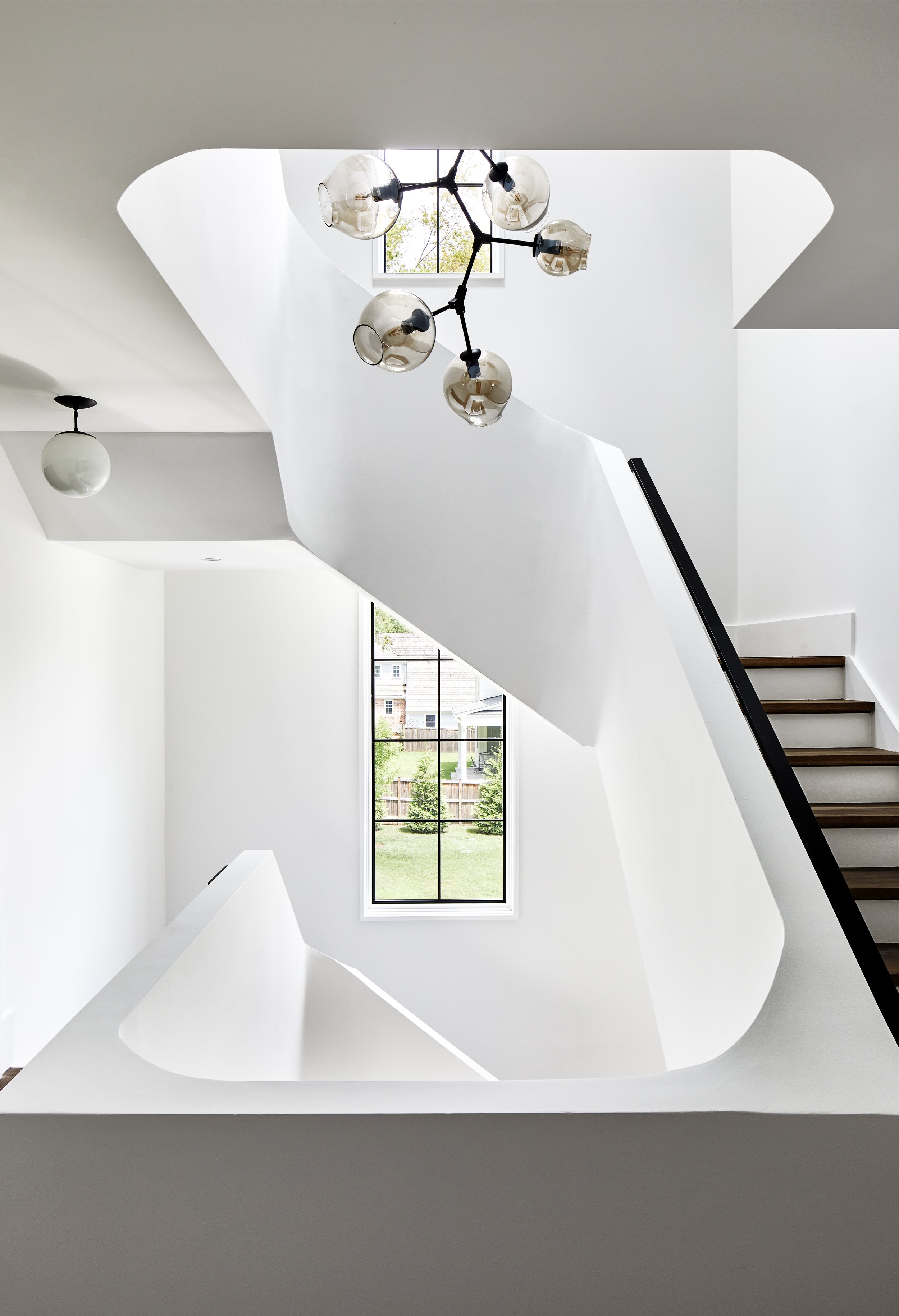 Modern staircase with black handrail, white walls, large window with a view of greenery outside, and contemporary ceiling light fixture with multiple globe-shaped bulbs.