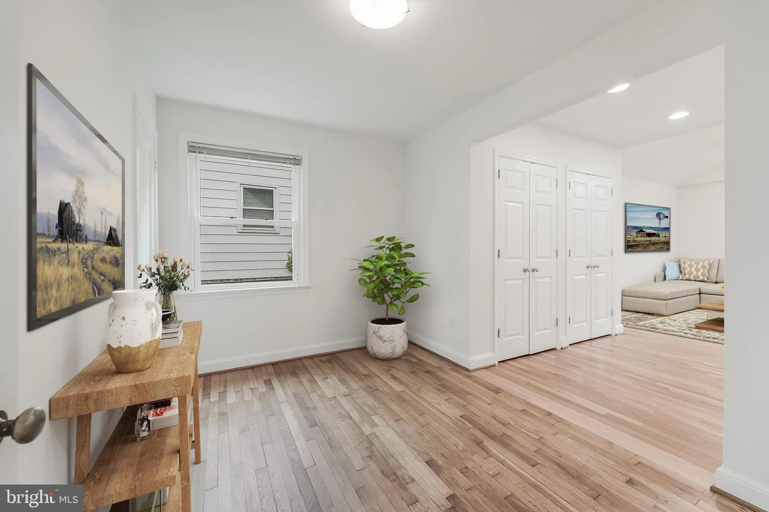 A clean, bright living room with hardwood floors, white walls, a window with blinds, a small wooden table with a vase and flowers, a potted plant, and a glimpse of a sitting area with a television and lounge chair.