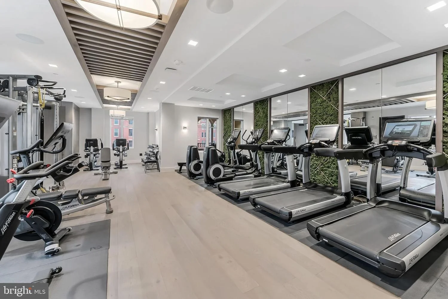 A modern indoor gym with various cardio and strength training equipment, including treadmills, ellipticals, stationary bikes, and weight machines, with large mirrors and windows letting in natural light.