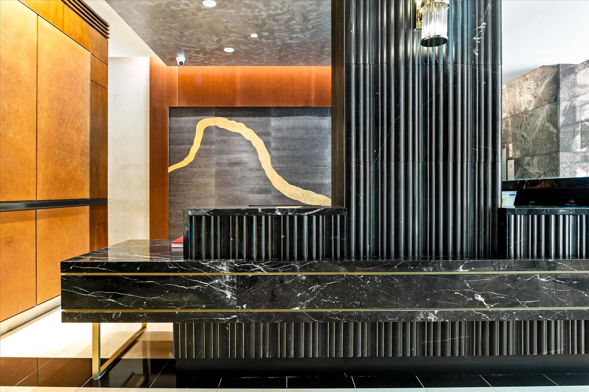 Modern hotel lobby reception desk with black marble surface and black ribbed marble front, in front of a patterned gray wall with a beige and yellow accent design, and a wooden wall with horizontal panels on the left. The ceiling has recessed lightin