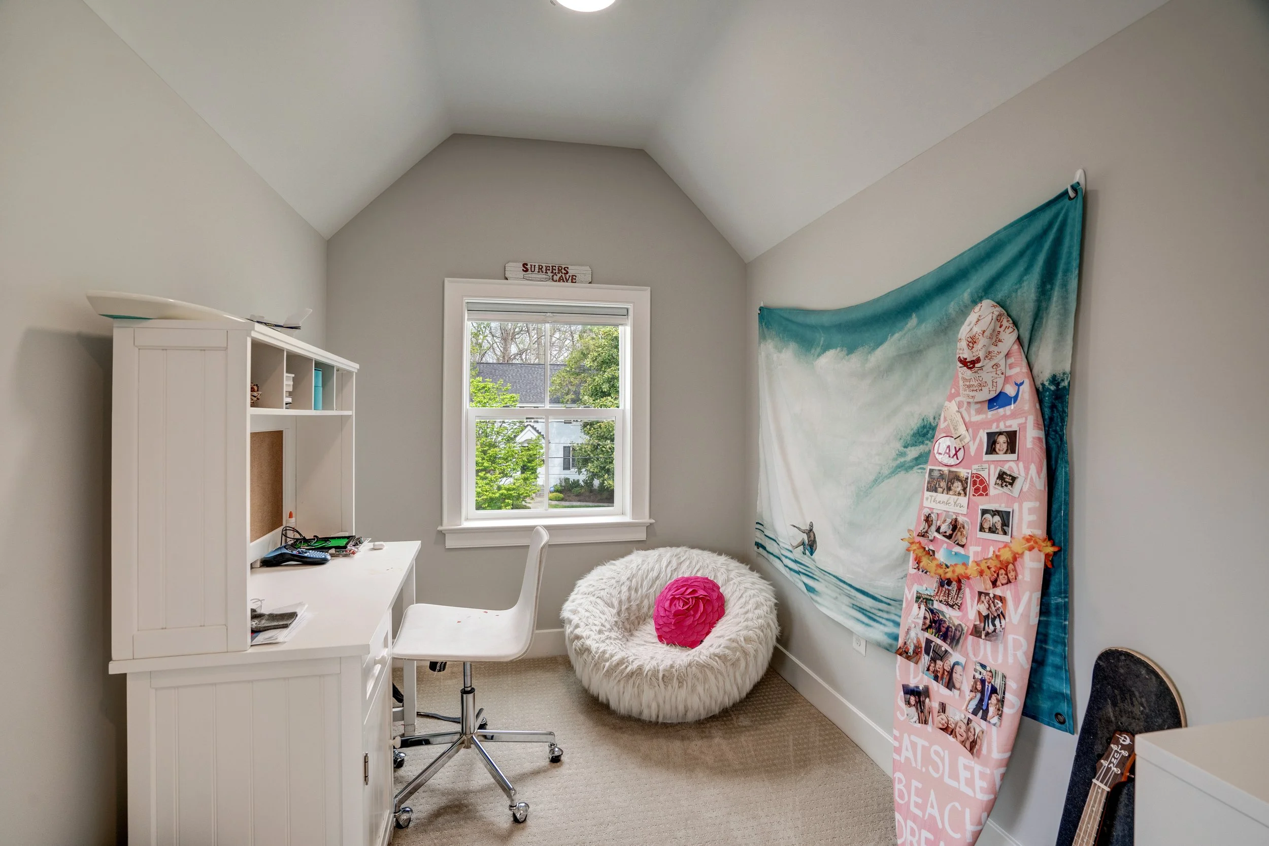 A small bright bedroom with a window, white desk, white fluffy chair, pink accent pillow, wall decor including a surf board sign, beach-themed tapestry, and photo collage.
