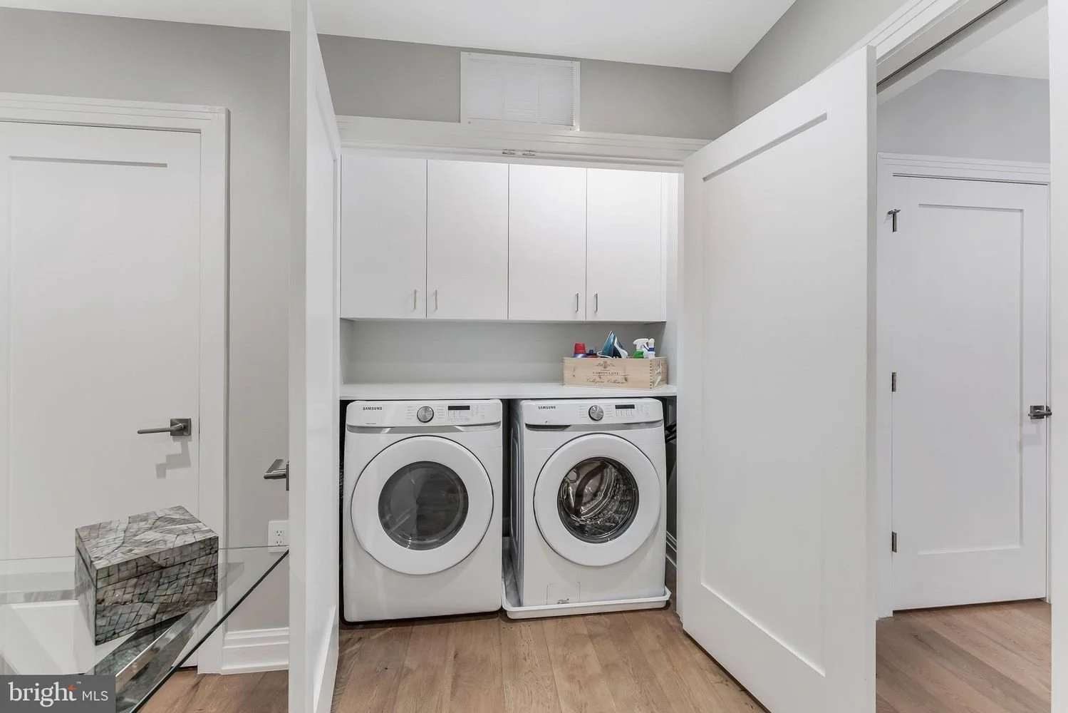 Laundry room with white washer and dryer, white cabinets above, and a wooden tray holding laundry supplies on top.