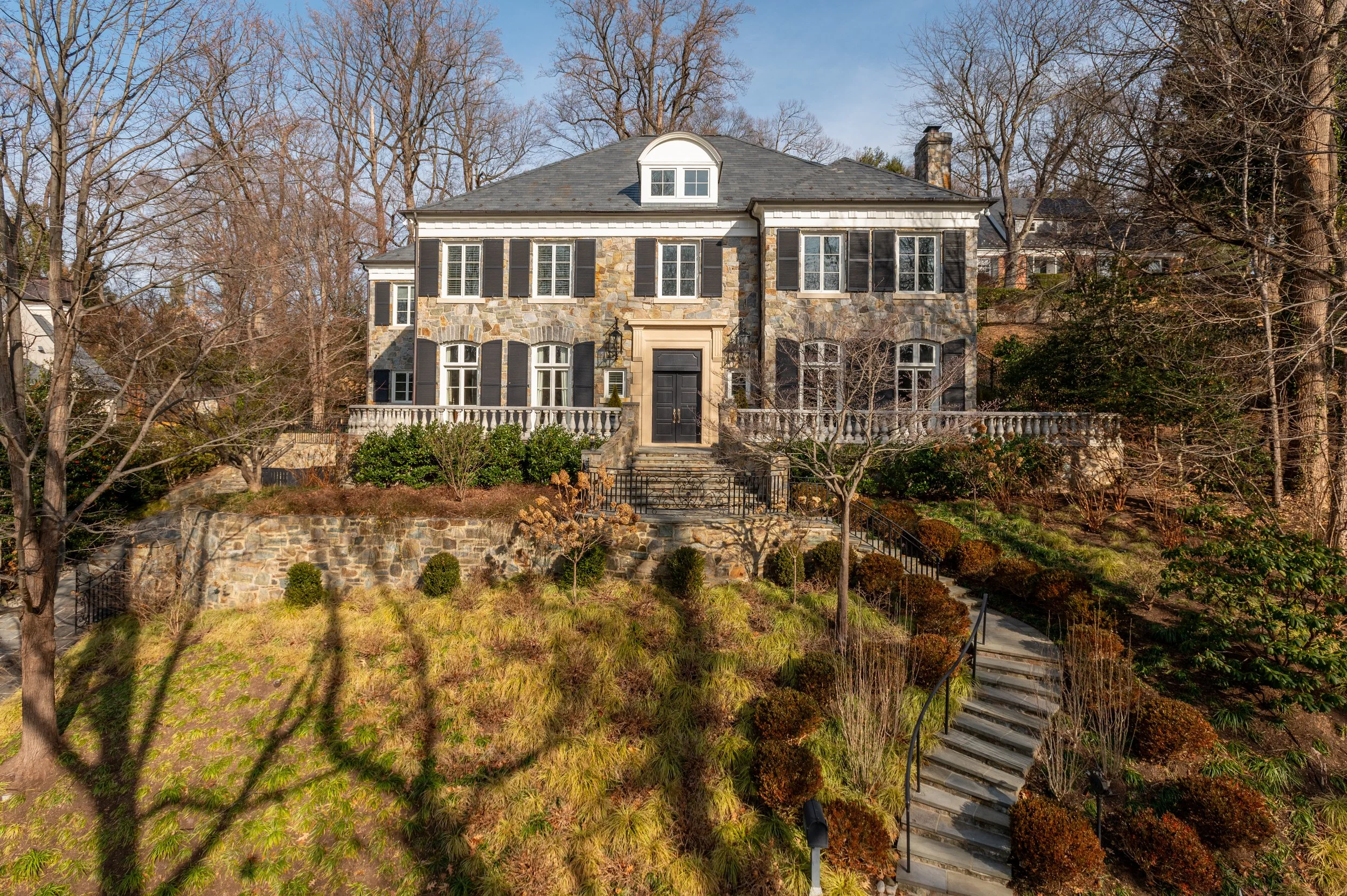 Front view of a large two-story house with a stone facade, black shutters, and a black front door, surrounded by leafless trees and landscaped yard with stairs leading up to the house.