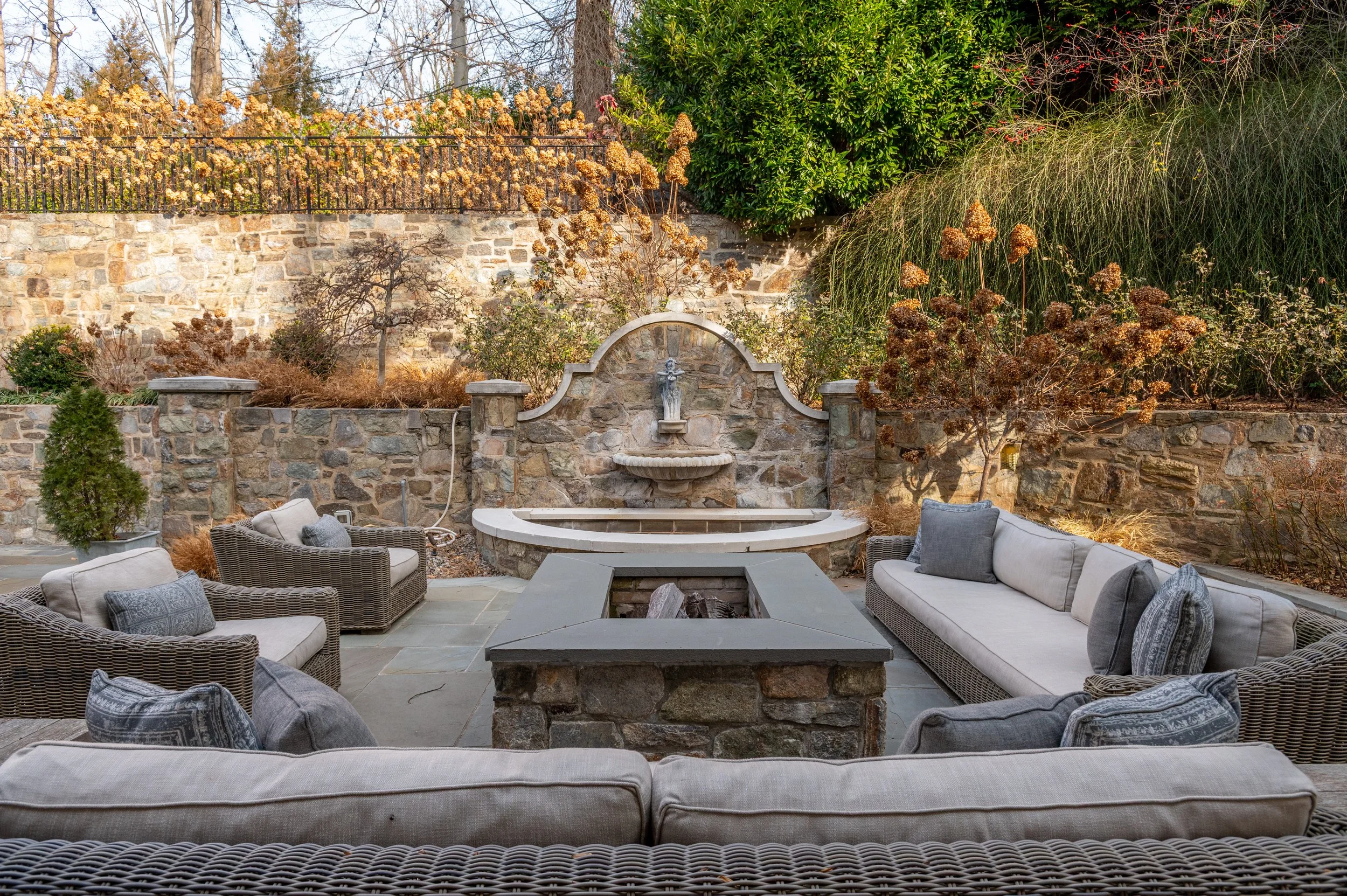 Outdoor patio with wicker seating and cushions, stone fountain, trees, bushes, and a brick and stone wall in the background.