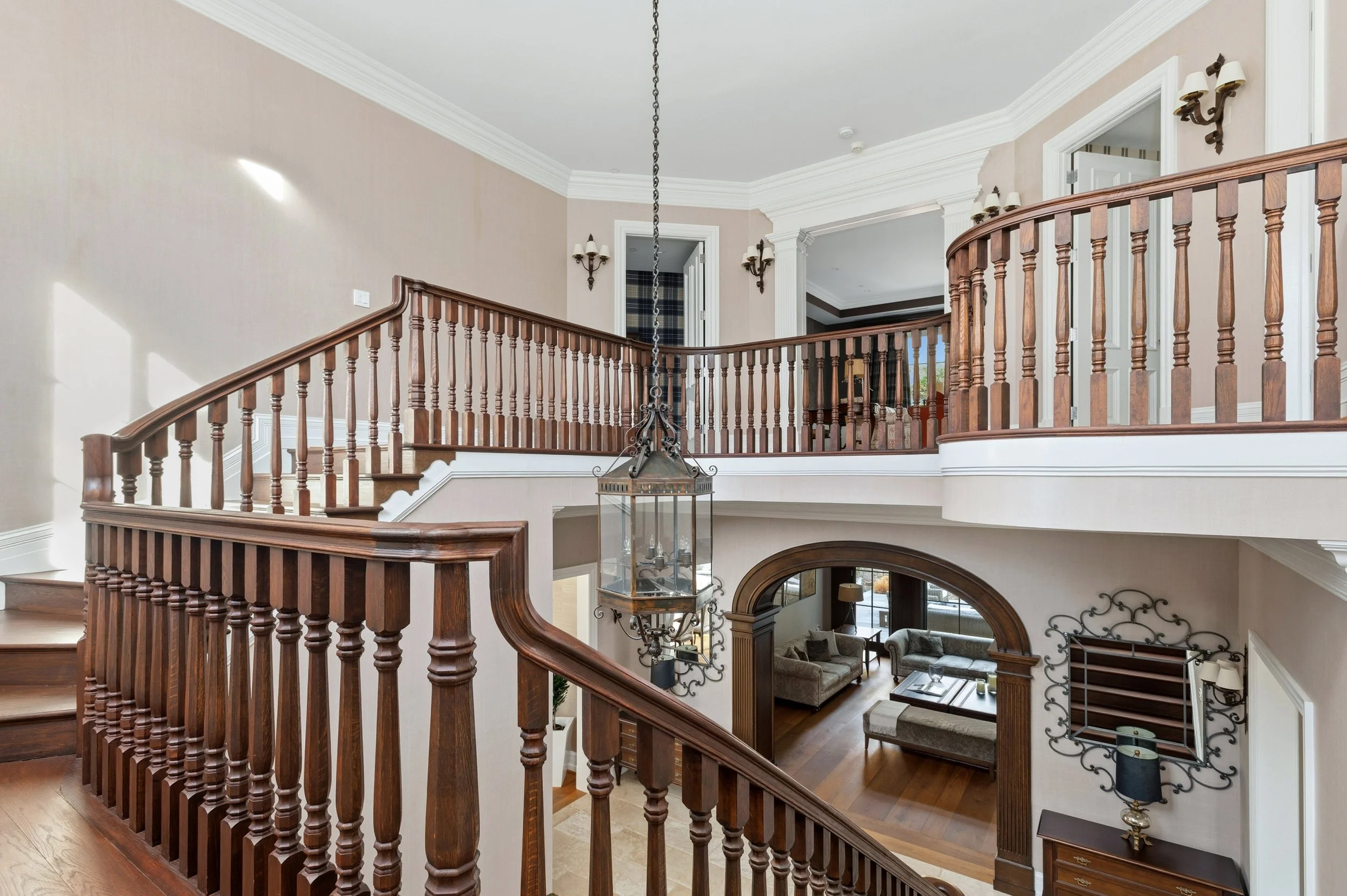 Interior view of a spacious multi-level home with a curved wooden staircase, chandelier, beige walls, and wall sconces in the bright living room.