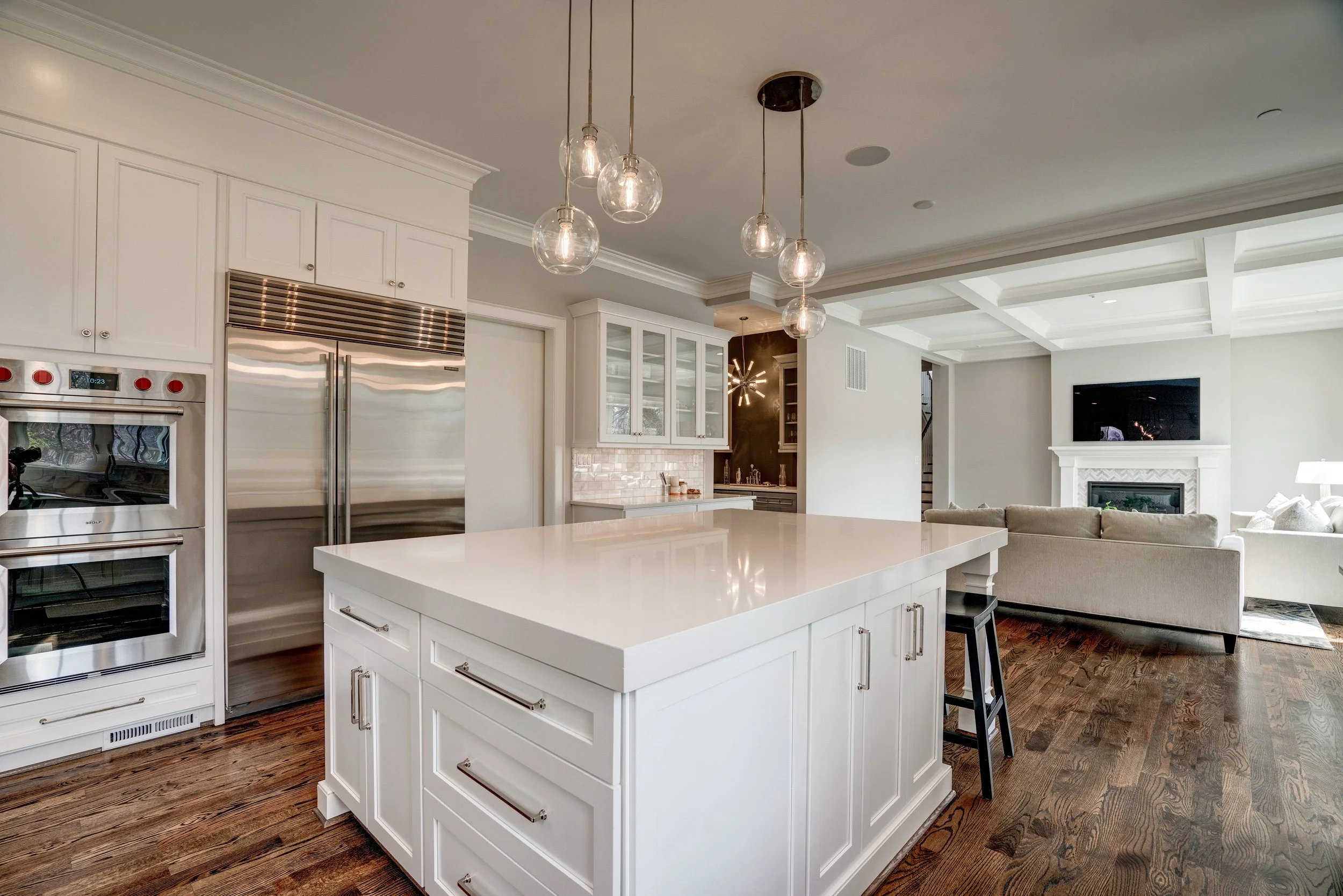 Modern open-concept kitchen with white cabinetry, a large kitchen island, stainless steel appliances, and a view into the living room with a fireplace and mounted TV.