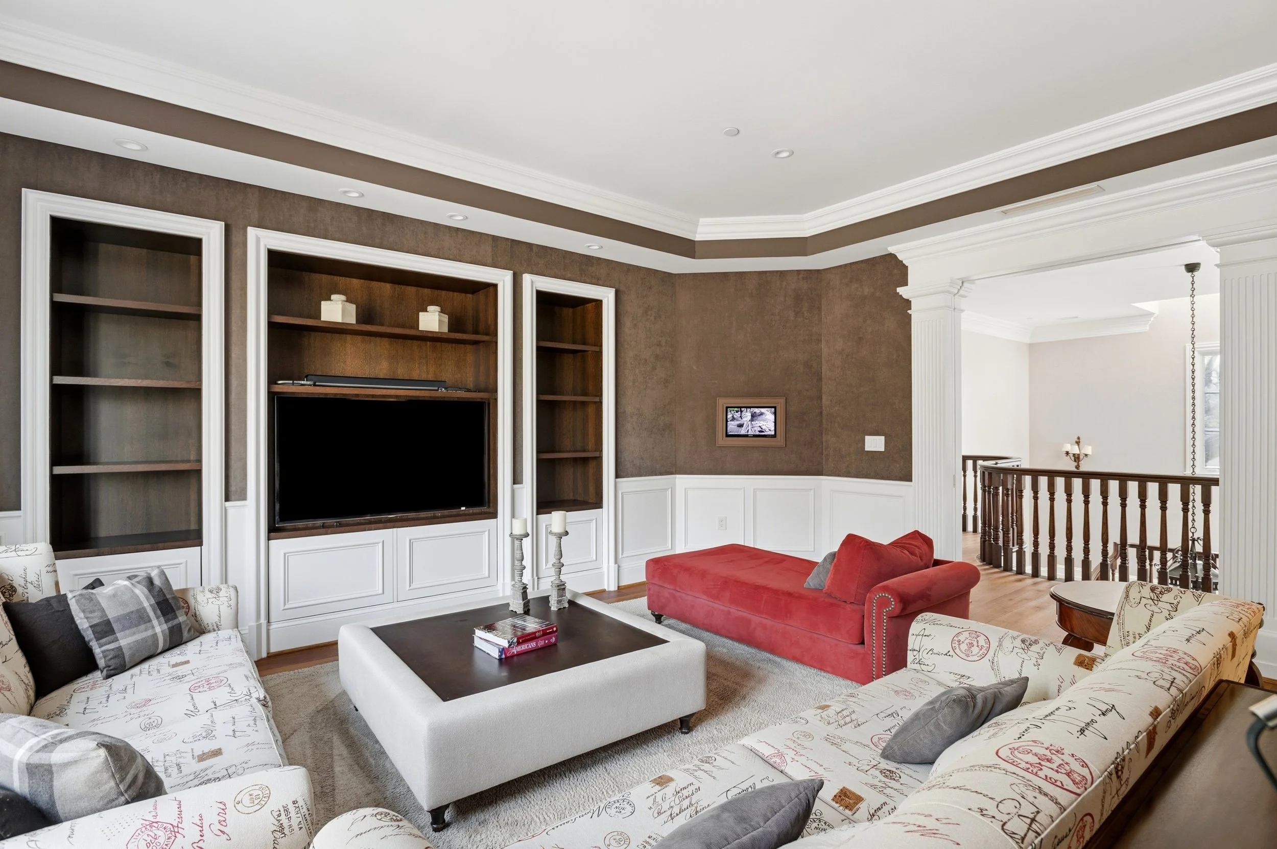 A living room with beige, white, and brown walls, white crown moulding, and a built-in wooden entertainment center. The room has a red upholstered ottoman, floral and checkered sofas, a white coffee table with books, and a small picture on the wall. 